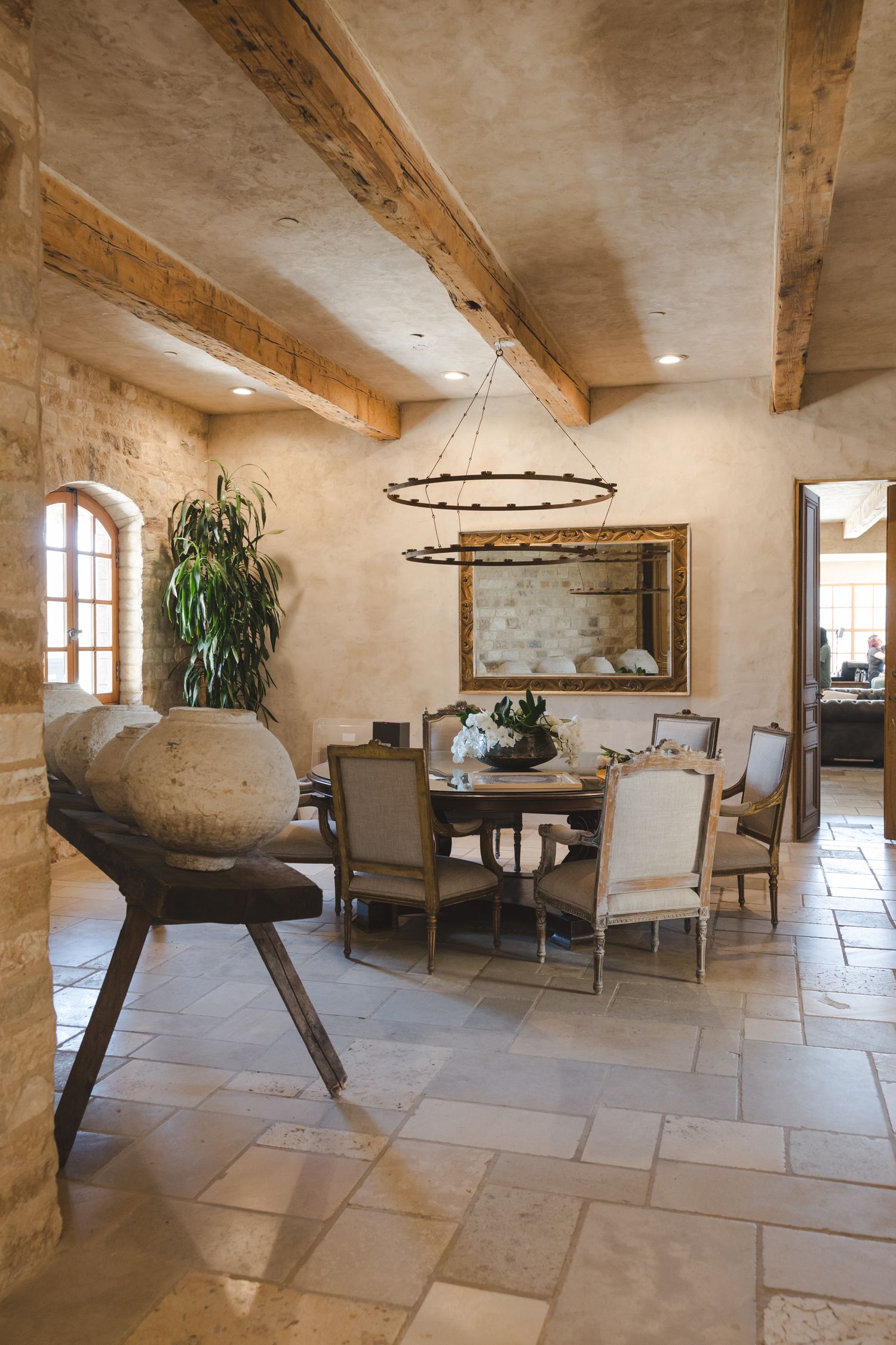 Rustic dining room with wooden beams, stone tile floors, upholstered chairs, and arched windows in a Mediterranean villa.