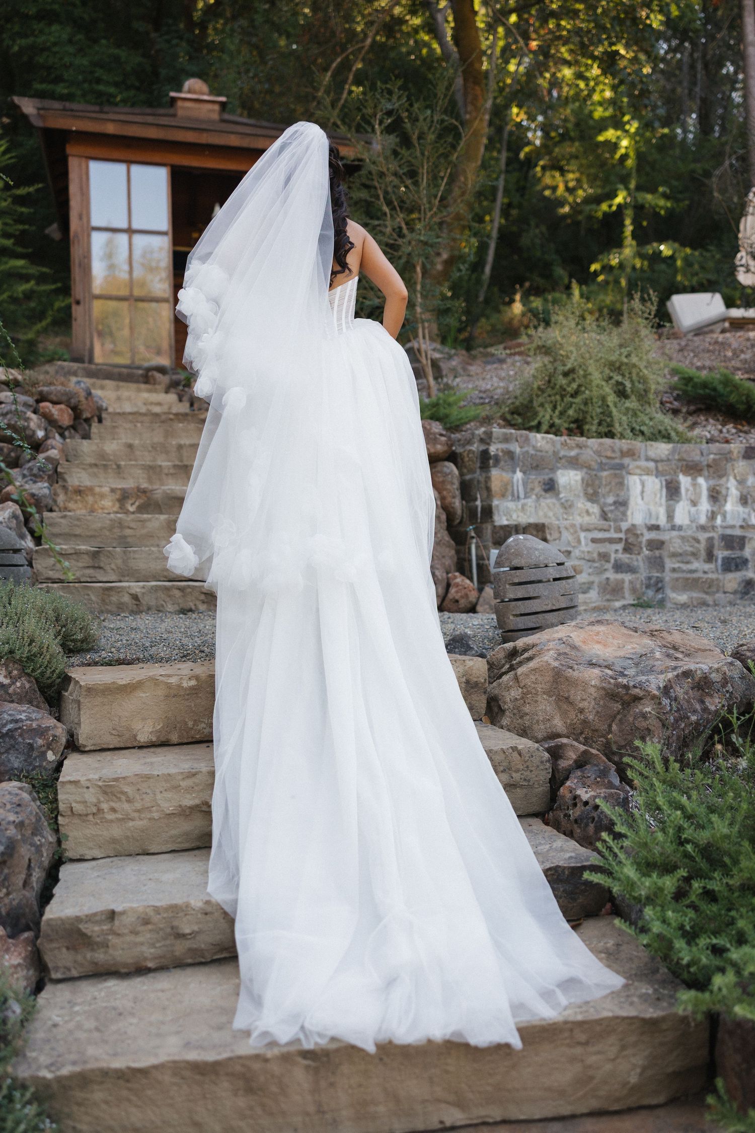 A flowing white wedding dress train cascades down stone garden steps in an outdoor setting.