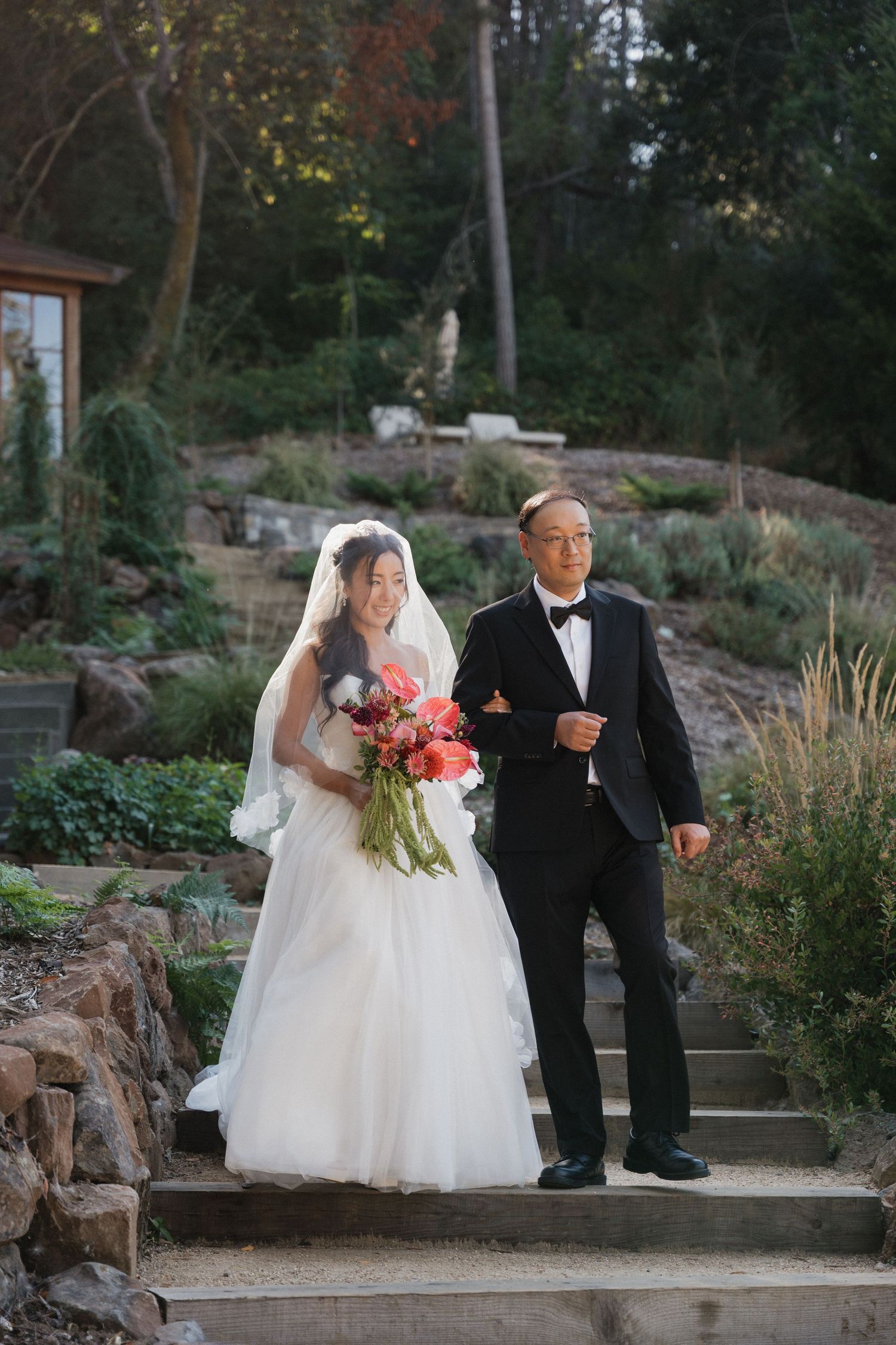 A bride in a white dress descends stone steps with her escort in a garden setting during a wedding ceremony.
