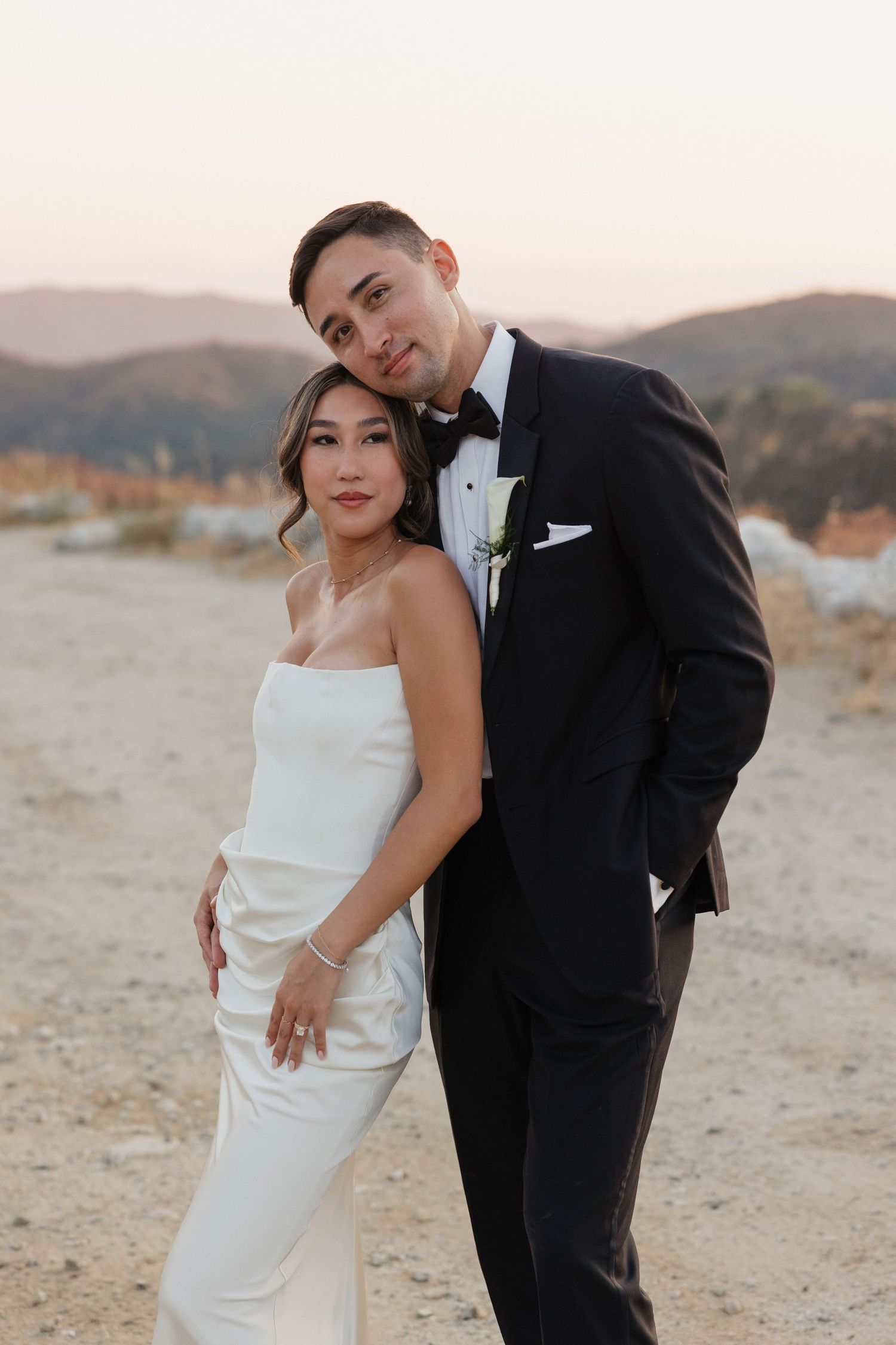 A couple in wedding attire poses romantically at sunset in a desert landscape with mountains in the background.