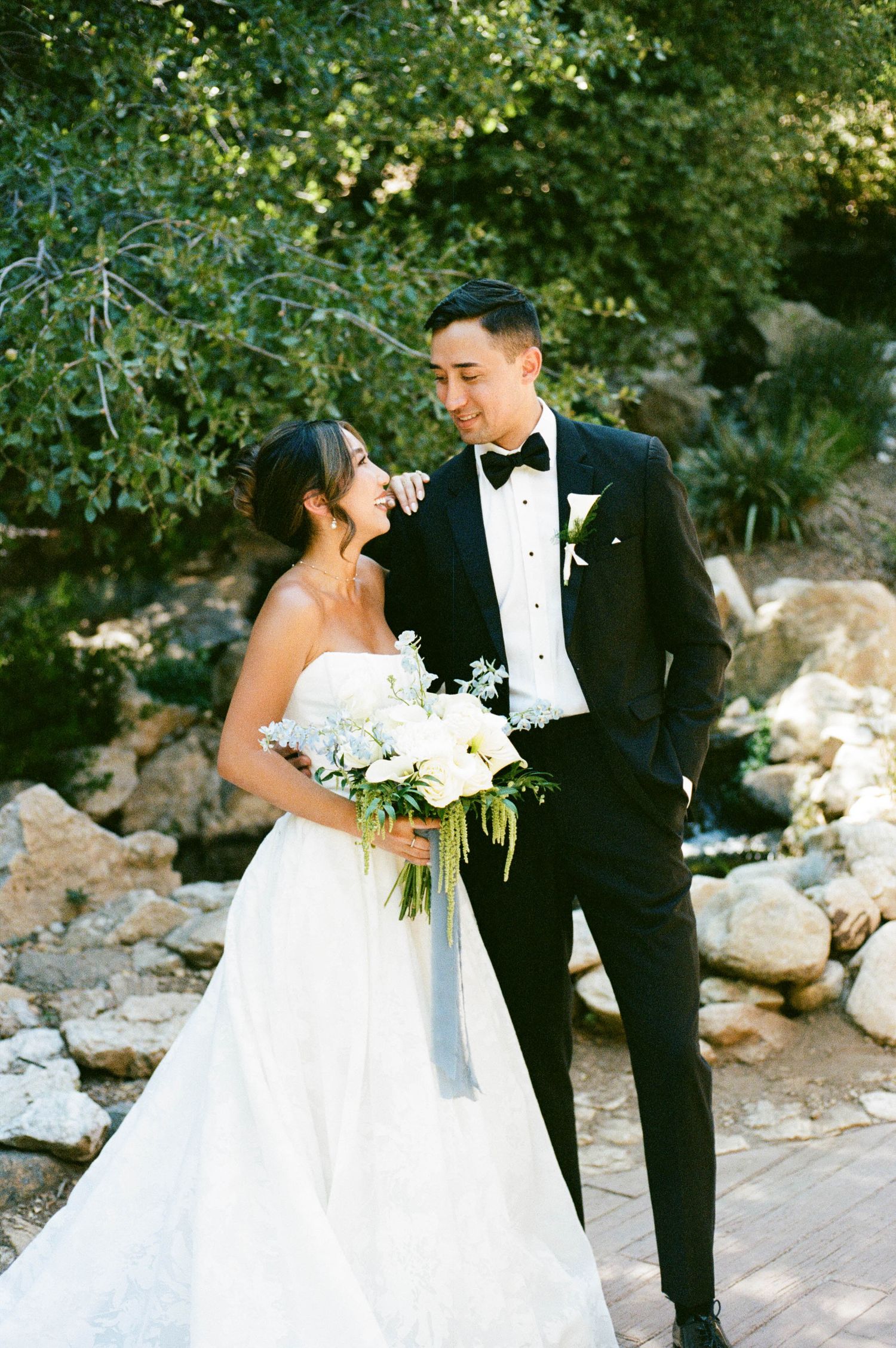 A newlywed couple poses for wedding photos in a natural setting with rocks and greenery.