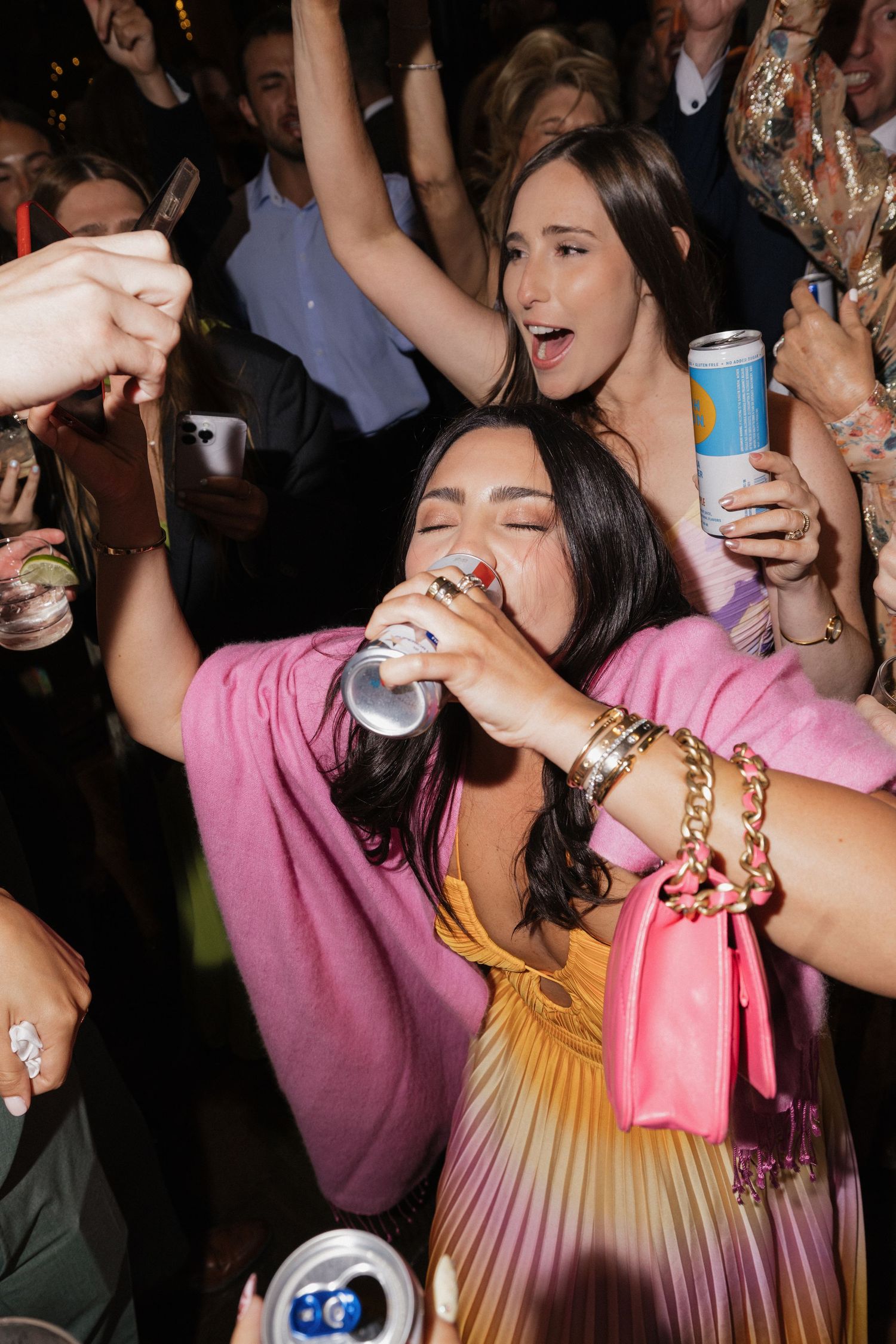 Woman in pink dress drinking from can while dancing at energetic wedding reception.