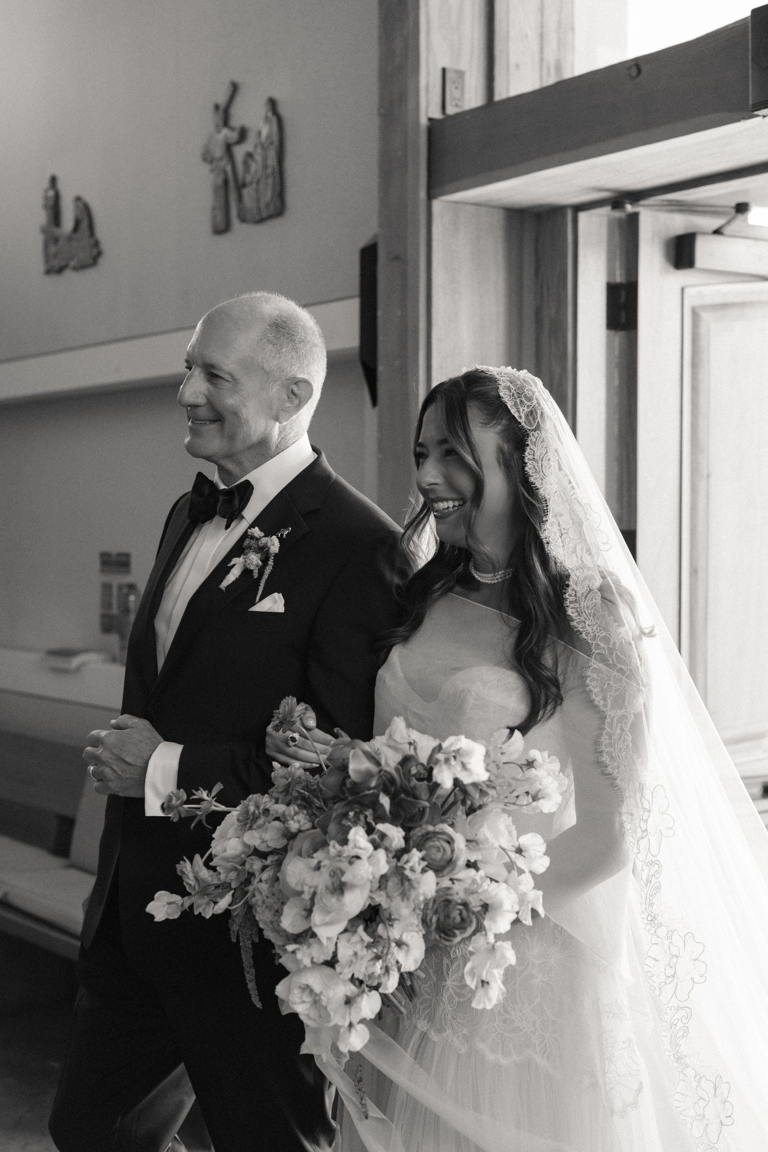 Black and white photo of smiling bride with lace veil and bouquet walking down aisle with older man.