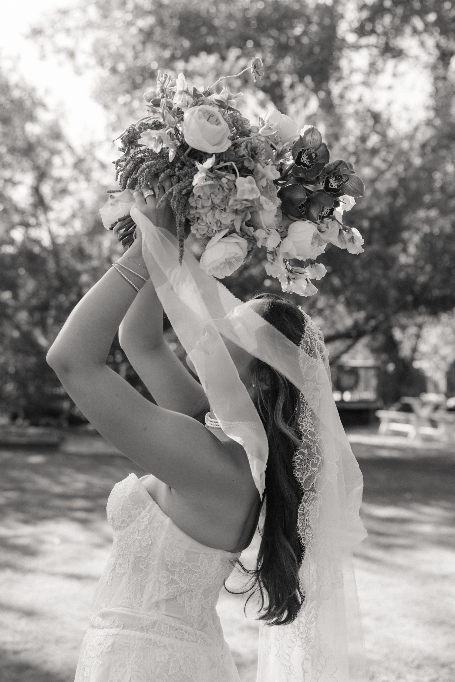 Black and white photo of bride in lace veil holding large floral bouquet overhead in sunlit garden.