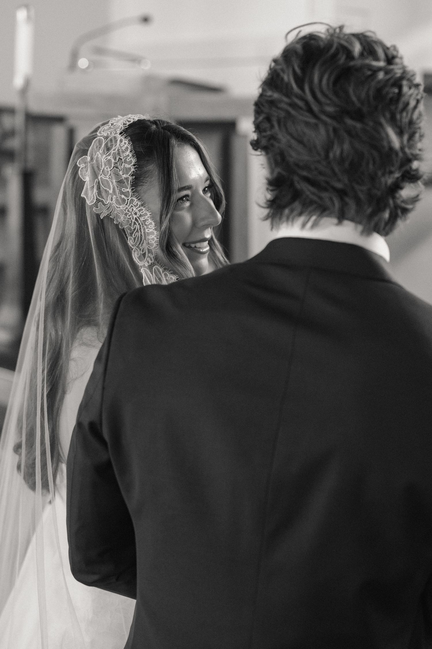 Black and white photo of smiling bride in lace veil exchanging vows with groom at altar.