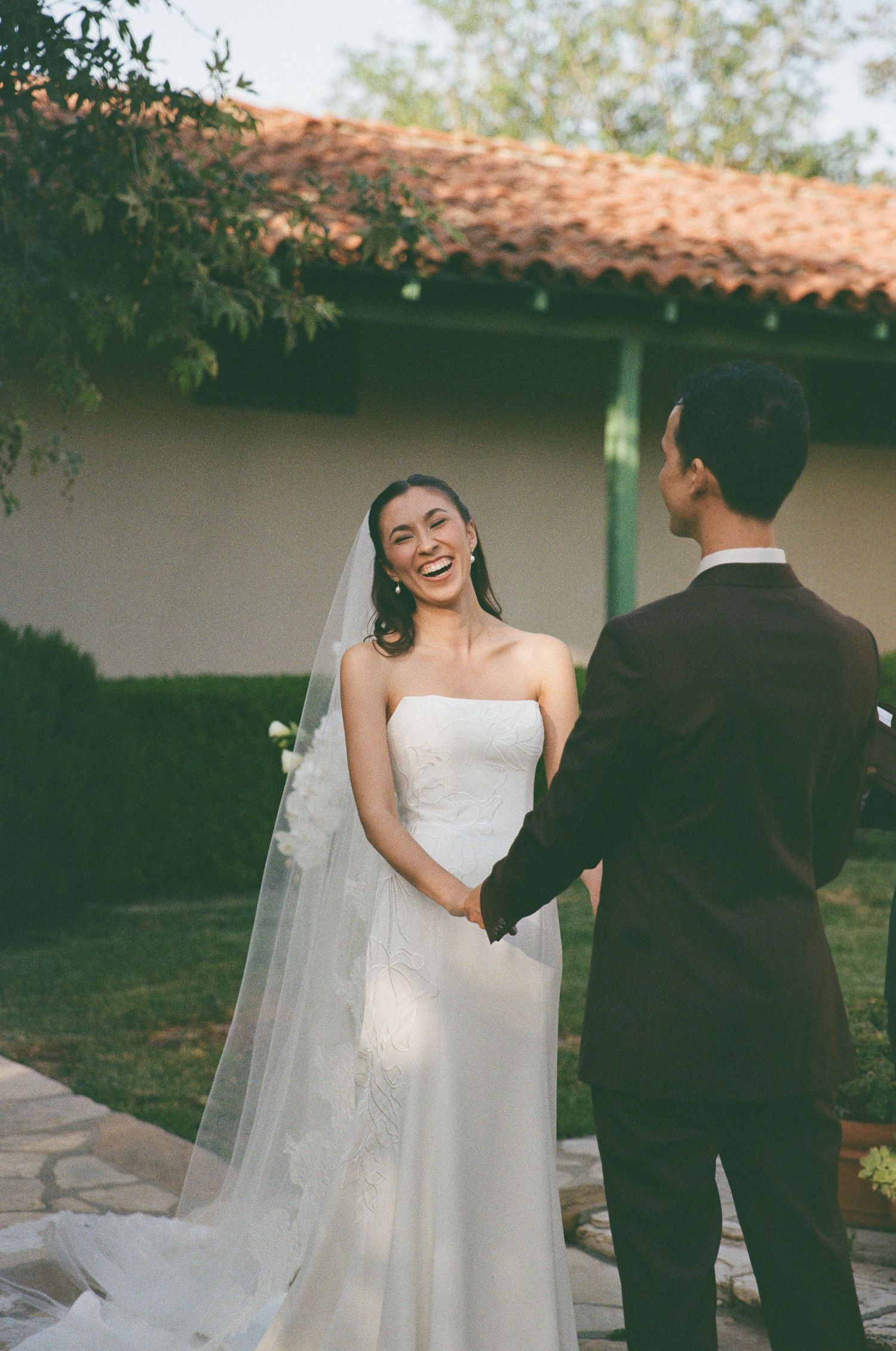 Smiling bride in white strapless gown and veil holds hands with groom in black suit outdoors at wedding ceremony.