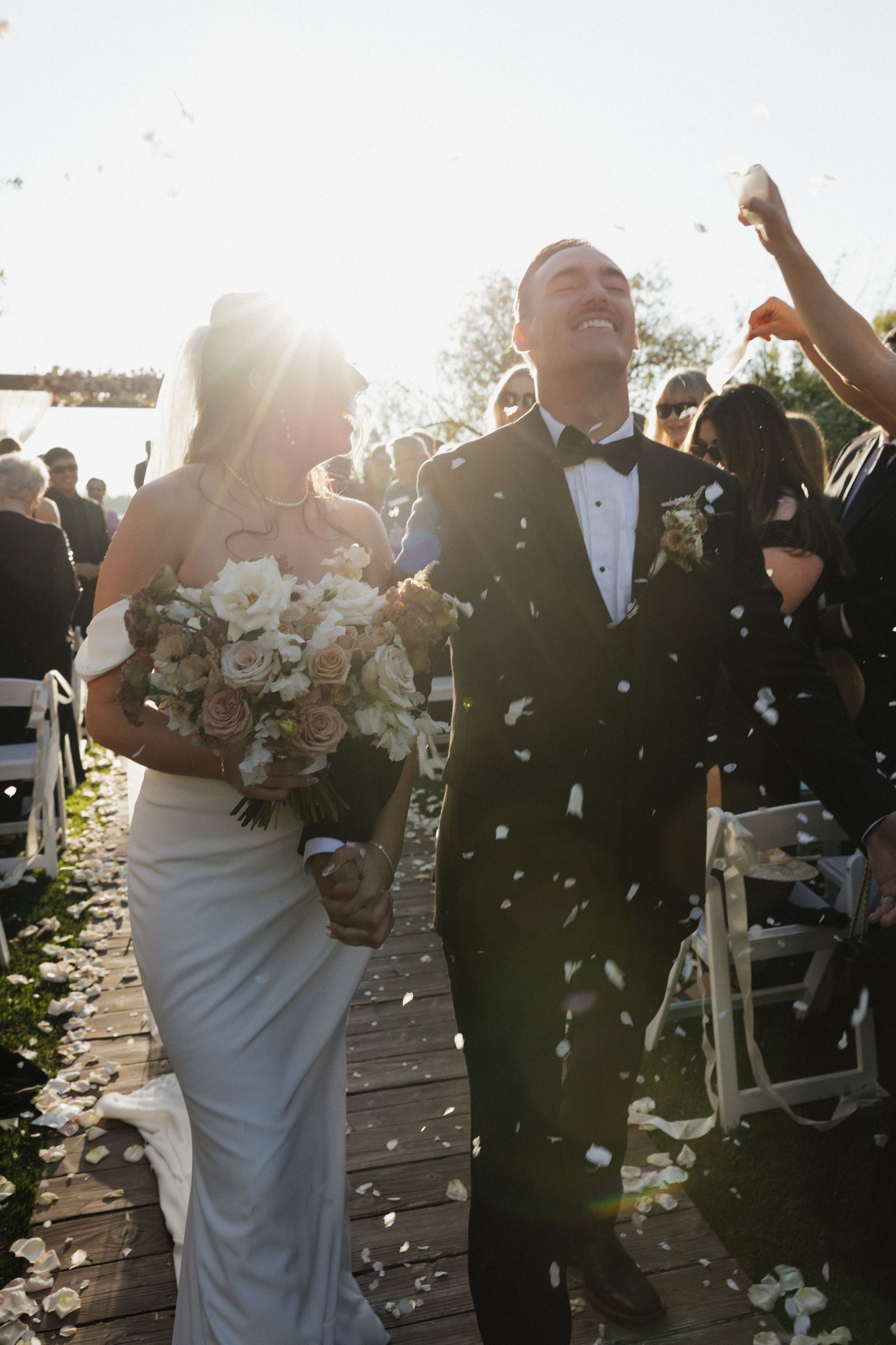Joyful newlyweds walk down the aisle as guests toss white flower petals in a sunlit outdoor wedding ceremony.
