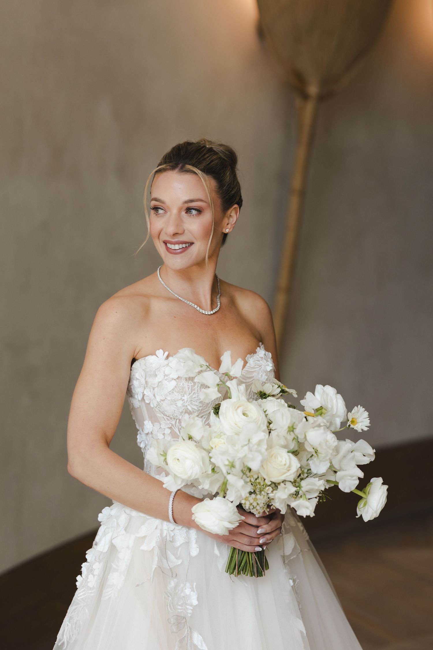 A bride in a strapless white lace wedding gown holds a luxurious bouquet of white flowers.