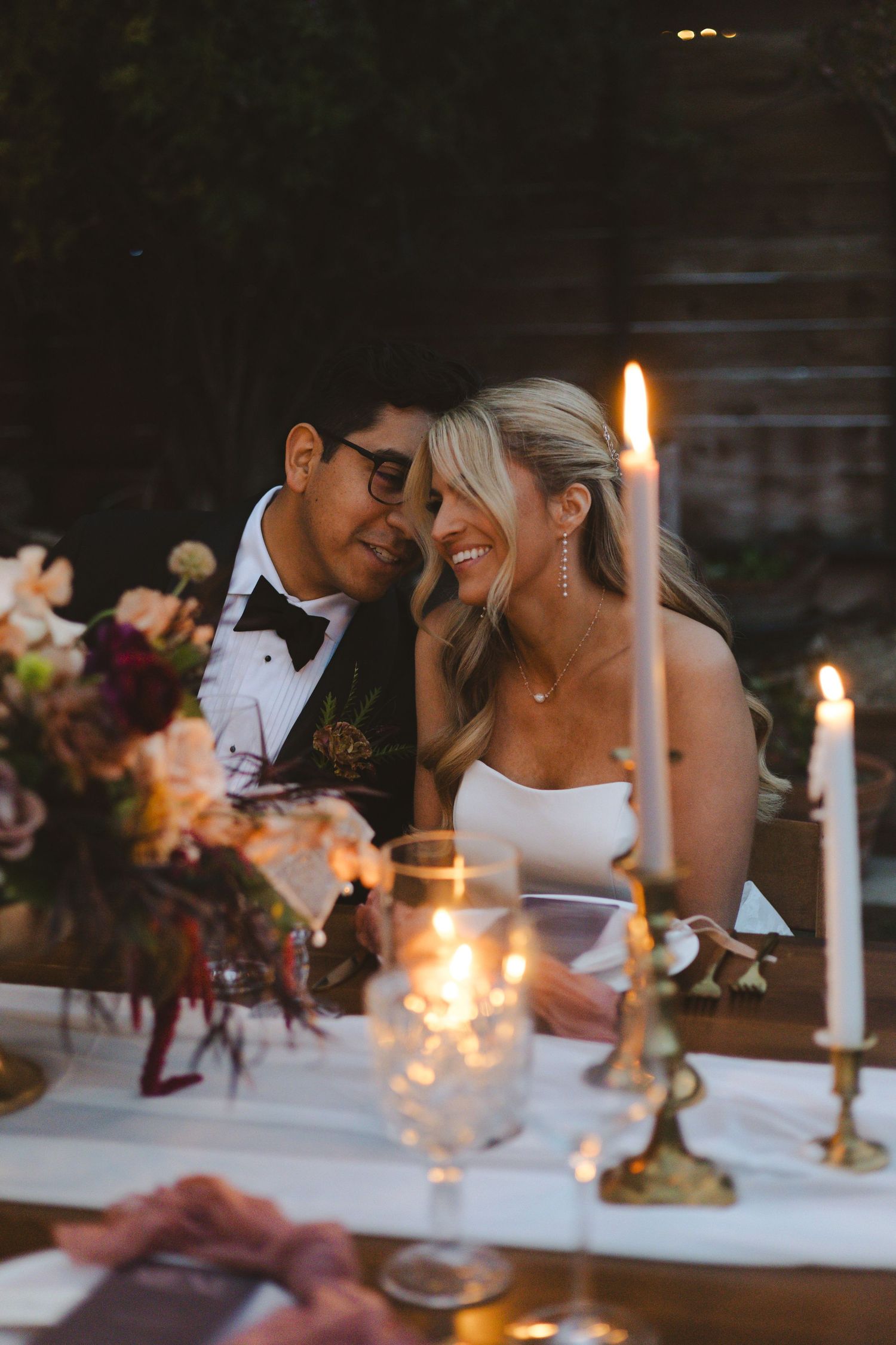 A romantic candlelit dinner scene with newlyweds sharing an intimate moment at an elegantly decorated table with flowers.