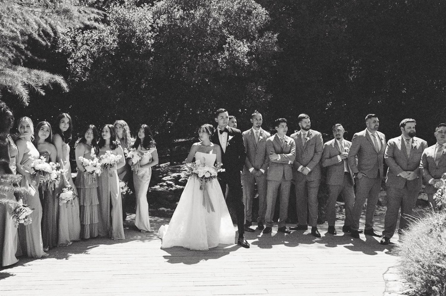 Wedding party lineup with bridesmaids in matching dresses and groomsmen in grey suits on garden path.