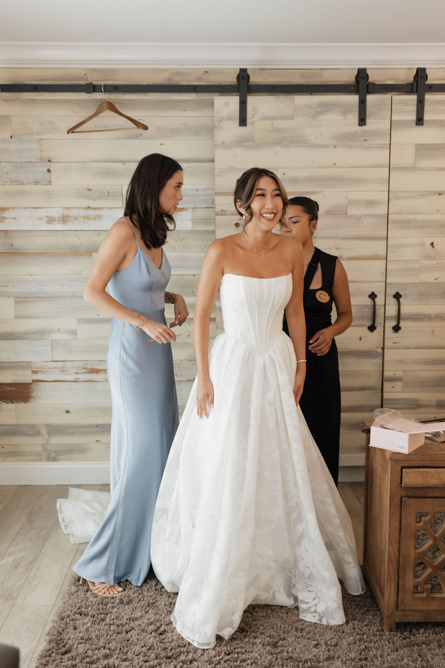 Bride in white gown getting ready with bridesmaids in a rustic room with wooden walls.