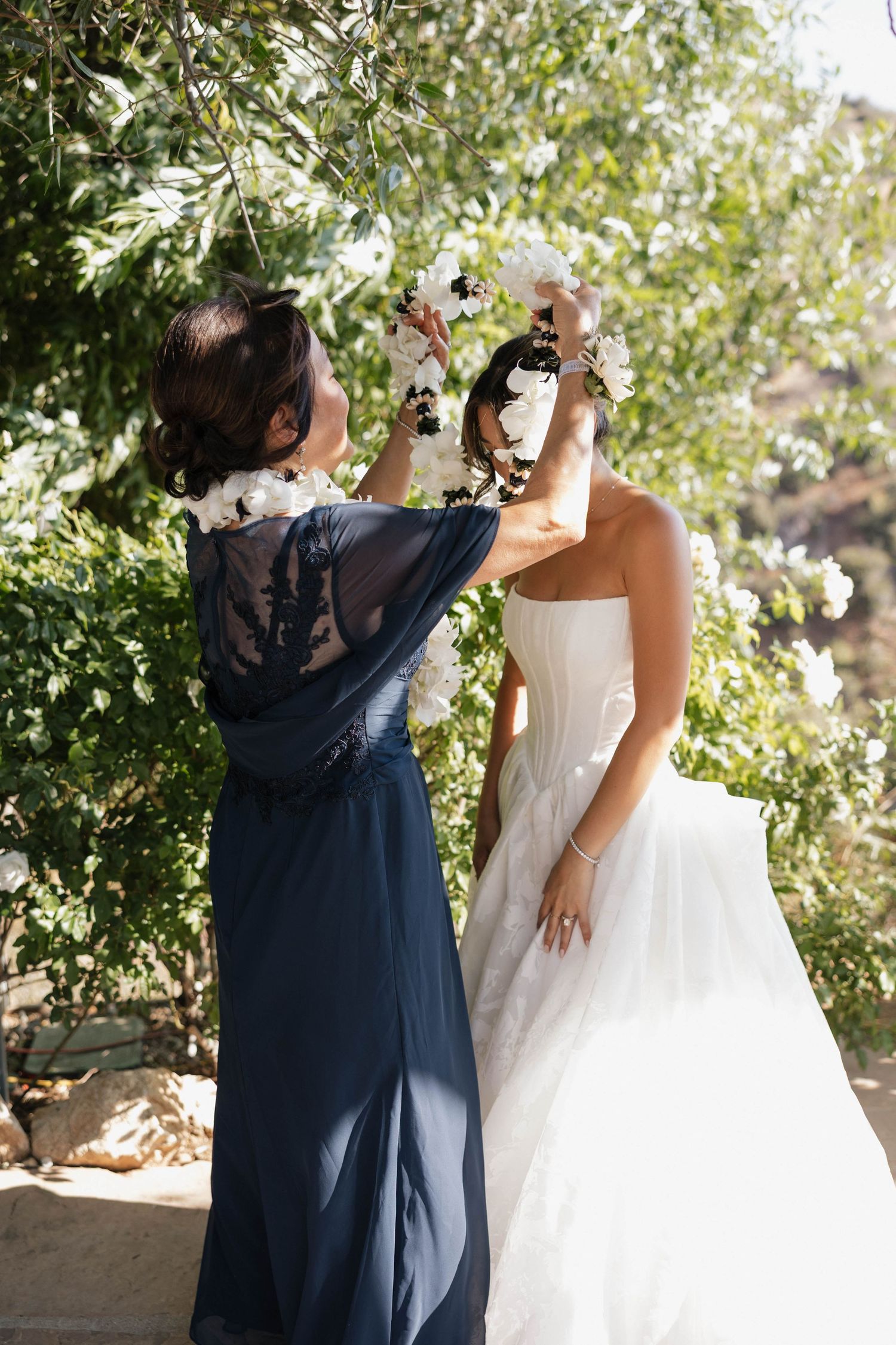A person in a navy blue dress helps adjust the wedding dress of another person in a garden setting.