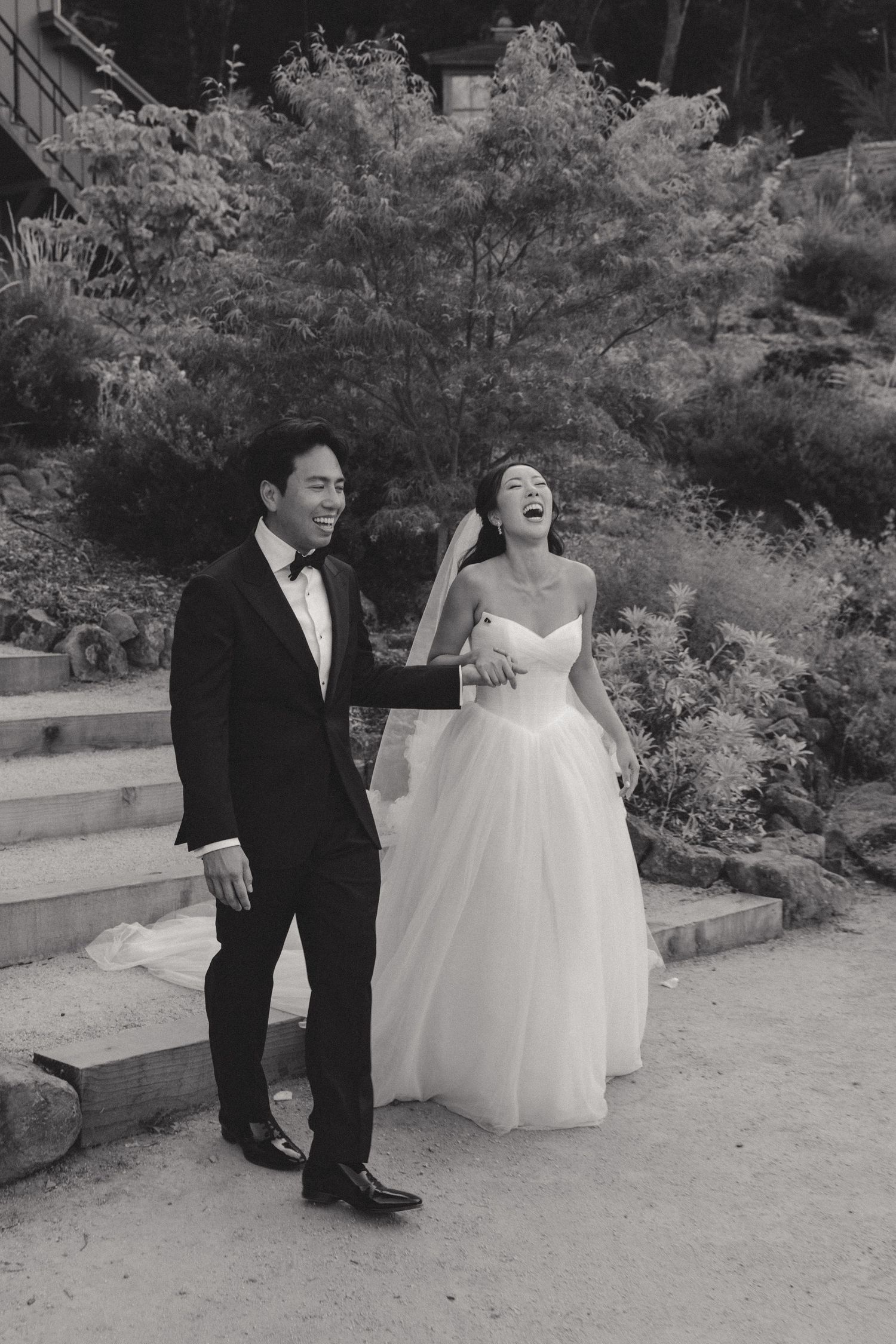 A couple in formal wedding attire stands together on a stone patio surrounded by gardens.