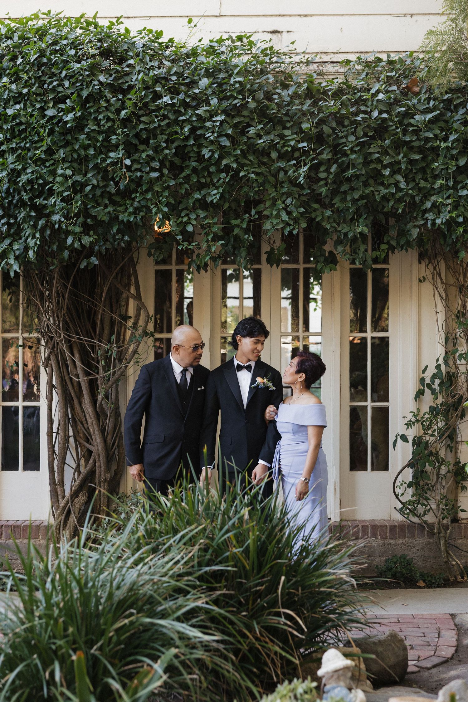 Wedding party gathered under an ivy-covered veranda with lush greenery and landscaping.