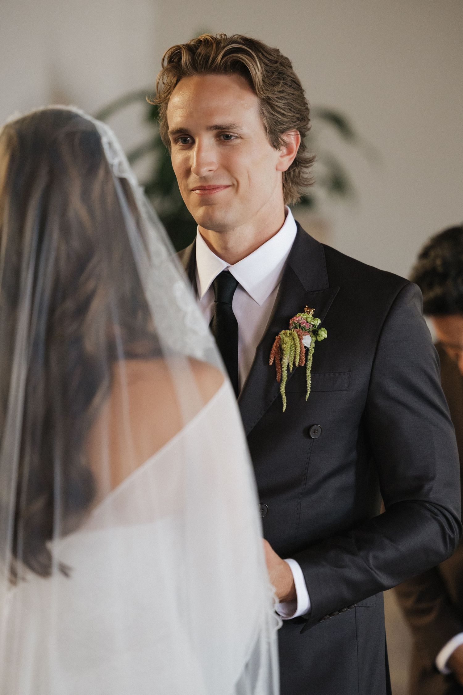Groom in black suit with floral boutonniere smiling at bride in wedding veil during ceremony.