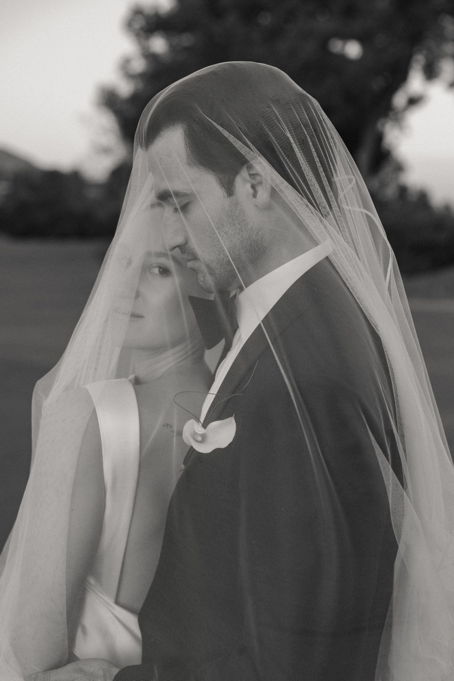 Black and white portrait of bride and groom sharing an intimate moment under a flowing wedding veil outdoors.