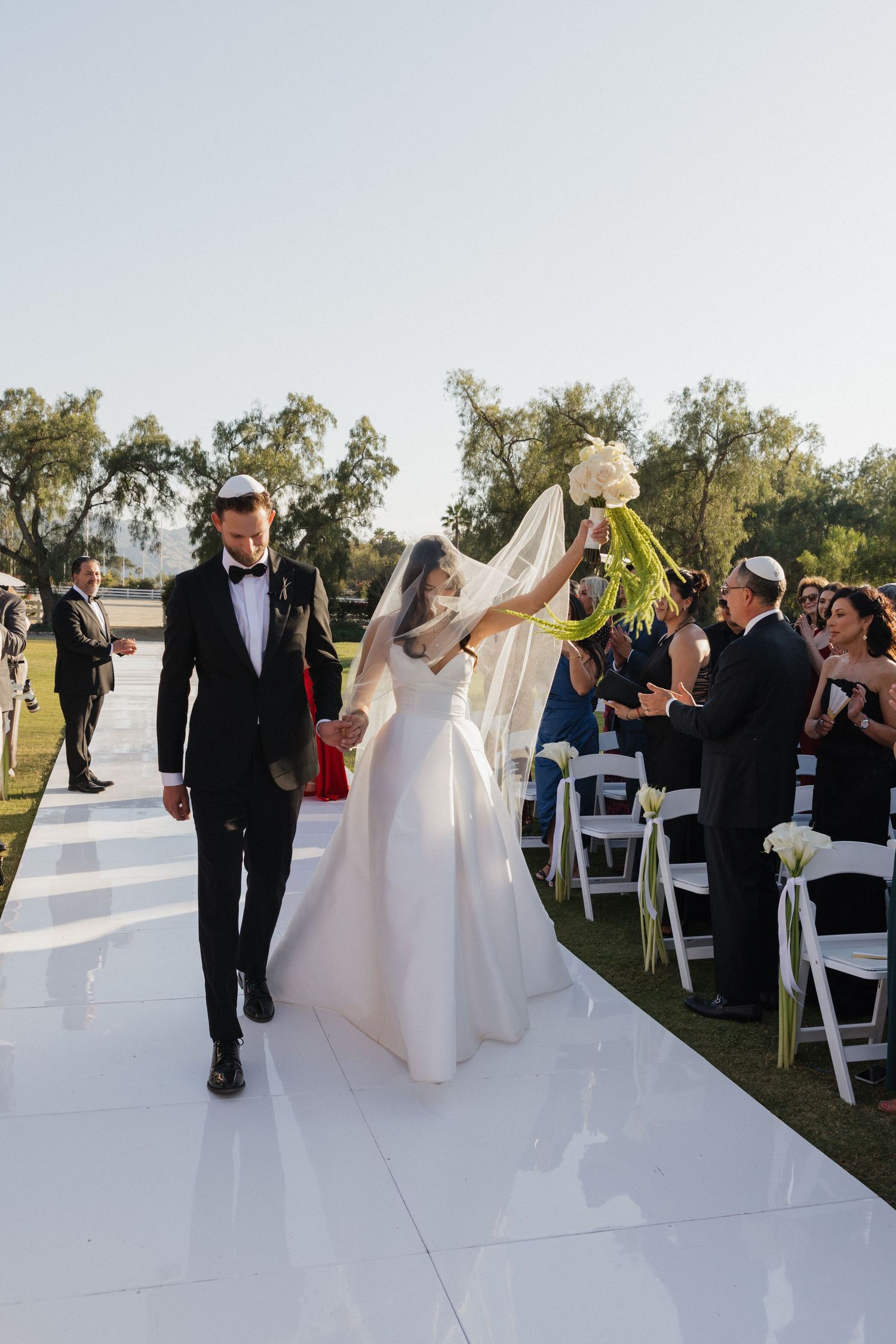 A wedding ceremony procession walks down an outdoor white aisle runner with guests seated in white chairs at sunset.