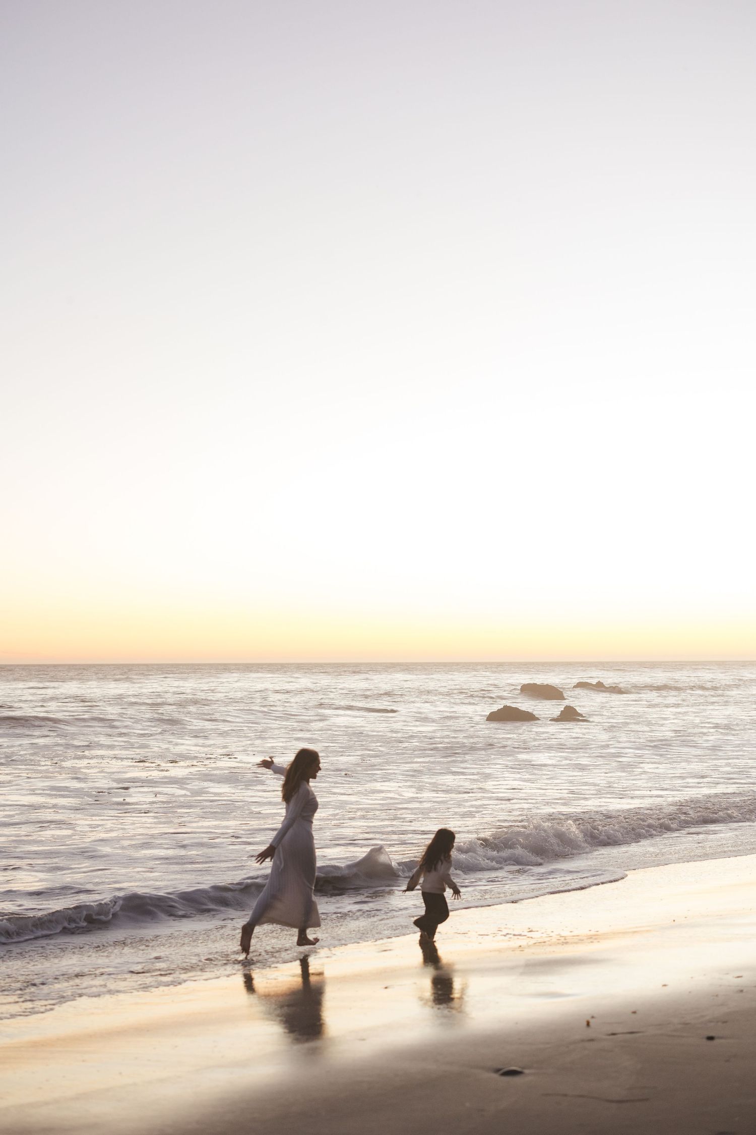 Two silhouettes walk along a serene beach at sunset with waves crashing against rocks in the distance.