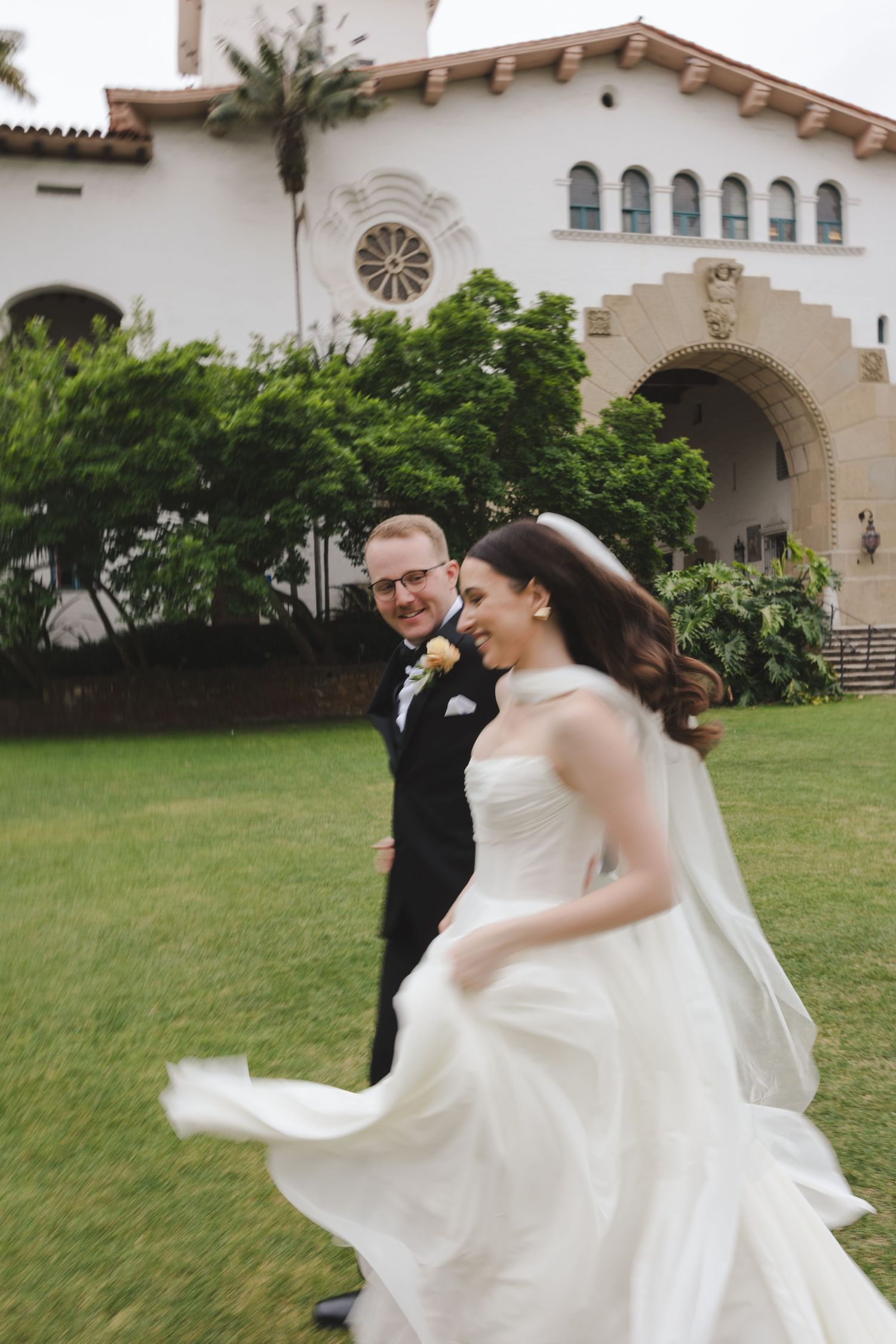 A couple shares a joyful moment on their wedding day outside a Spanish Colonial style church with white walls and arches.