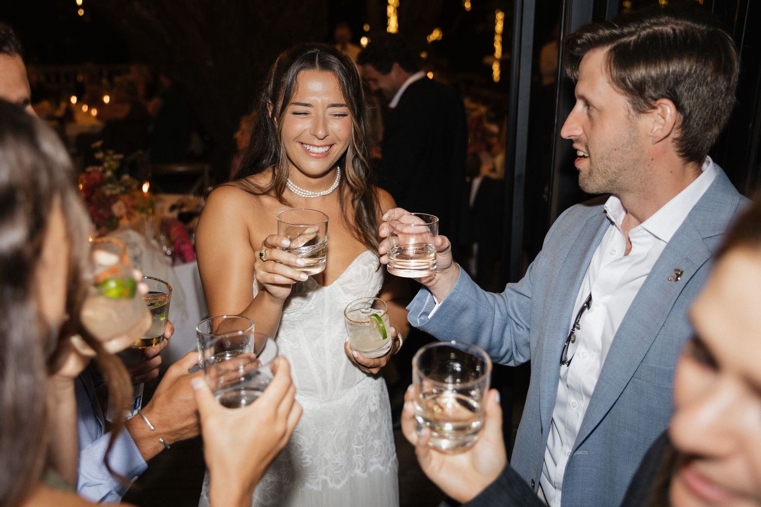 Bride and guests joyfully toast with cocktails during lively indoor wedding reception celebration.