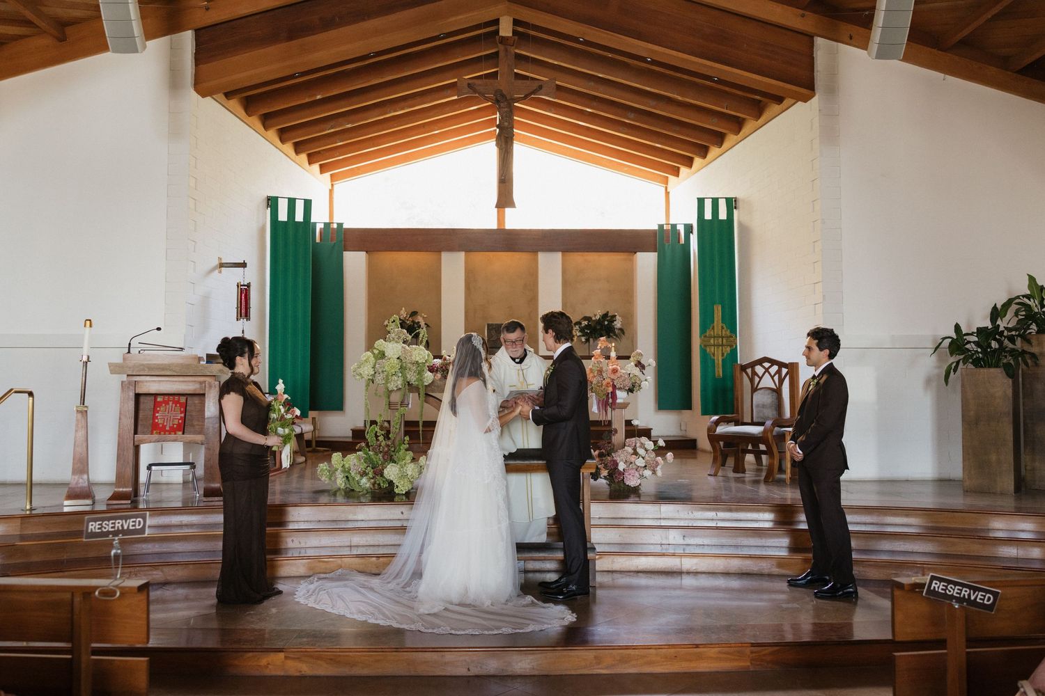 A bride and groom exchange vows at a church altar, flanked by wedding party members, with green banners and floral arrangements.