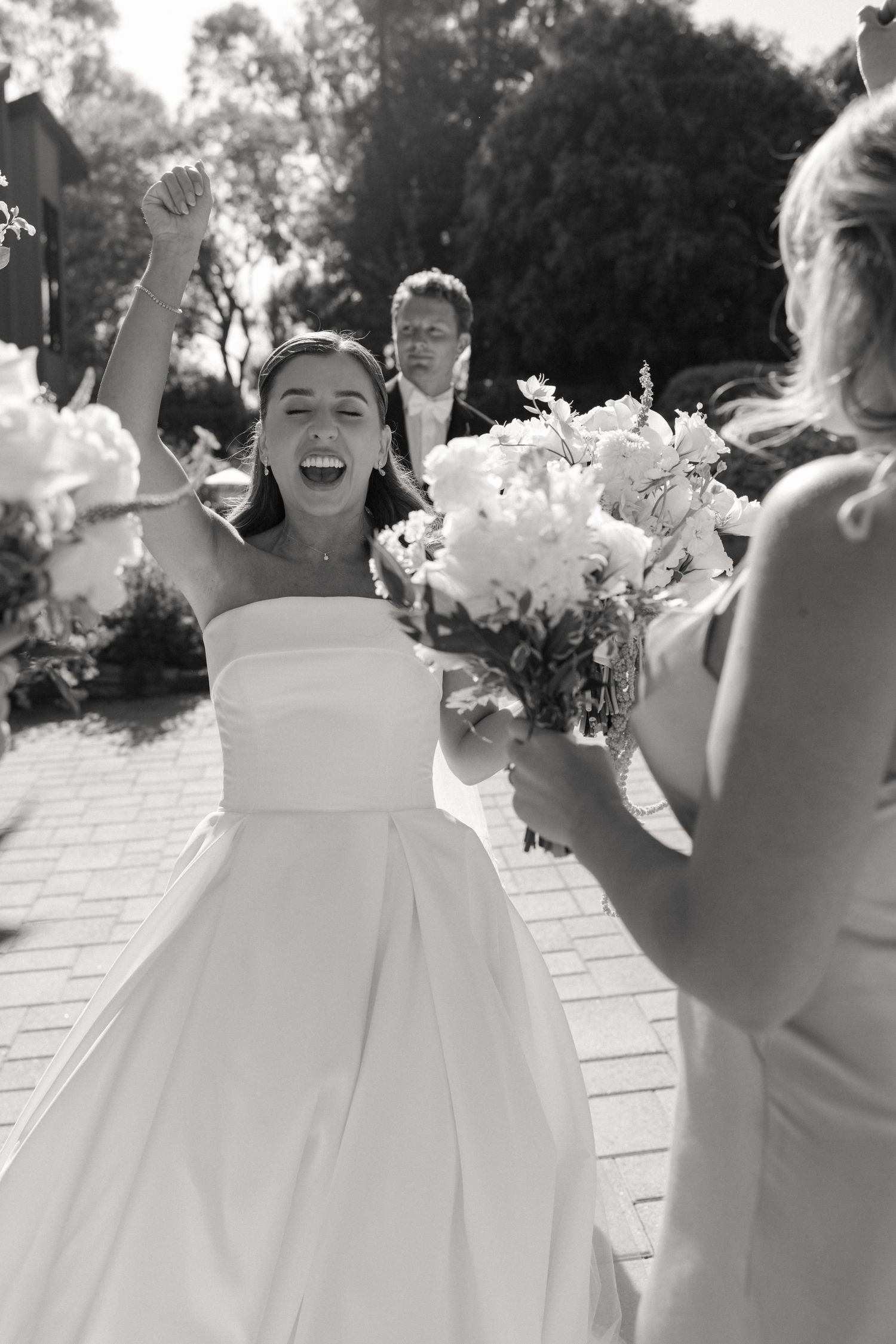 Black and white photo of joyful bride celebrating with raised fist, holding bouquet, groom smiling behind her.