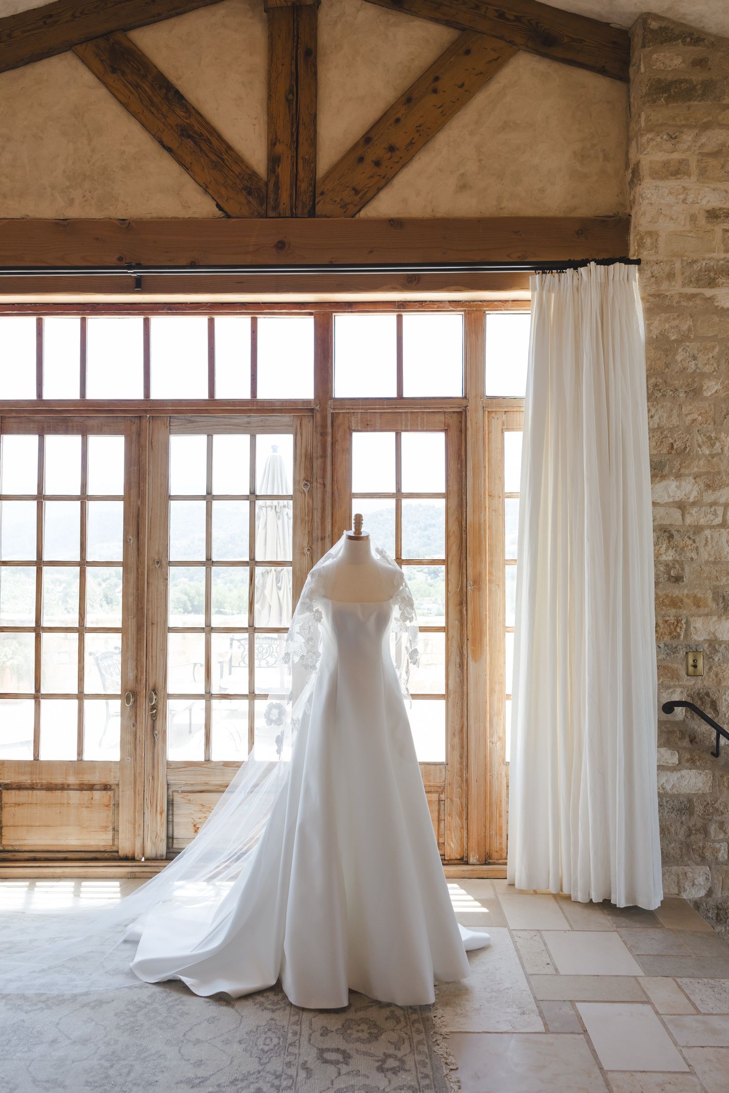 Elegant white wedding gown displayed in rustic room with wooden beams and large windows.