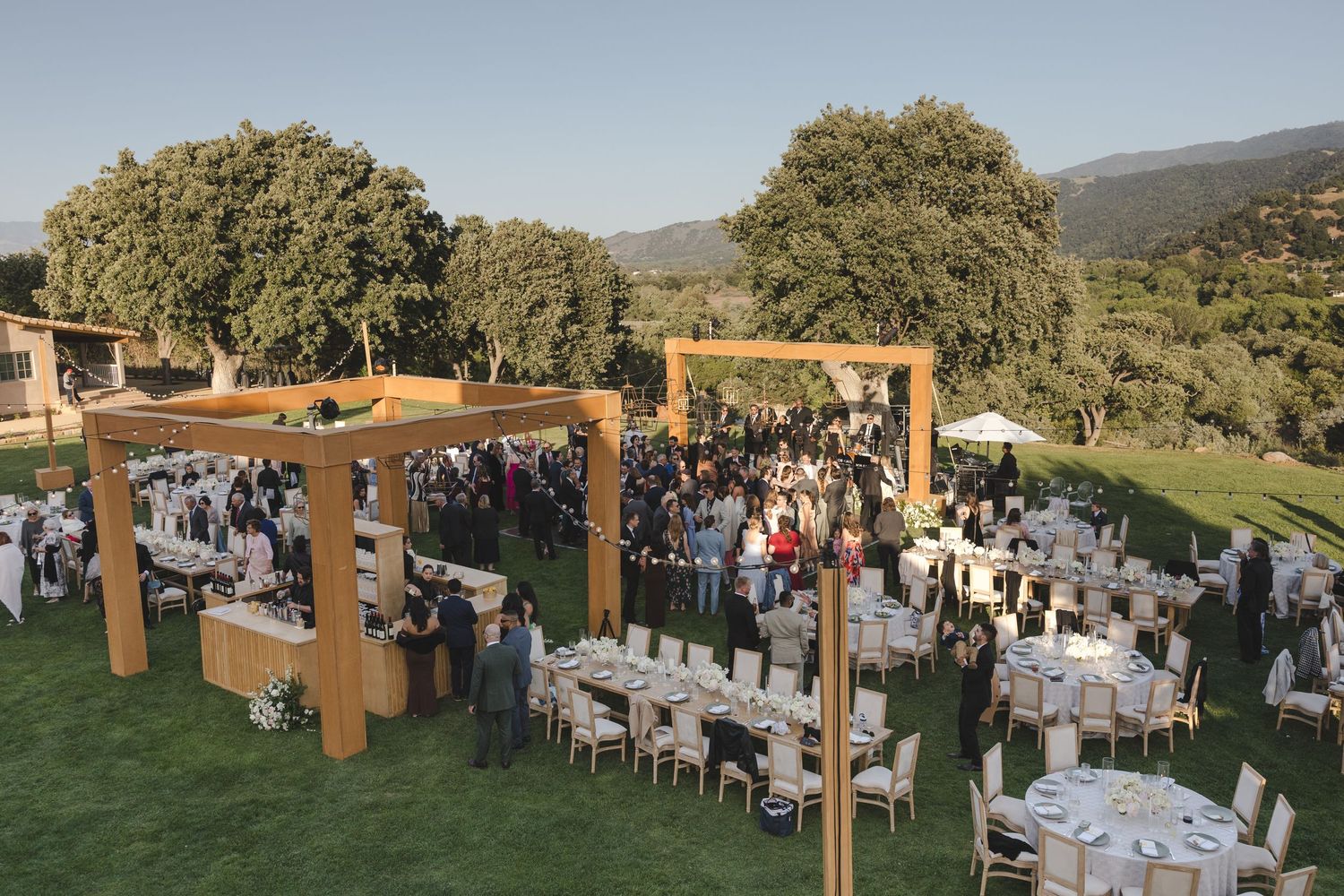Outdoor wedding reception setup with wooden pergolas and white table settings on a grassy hillside venue.
