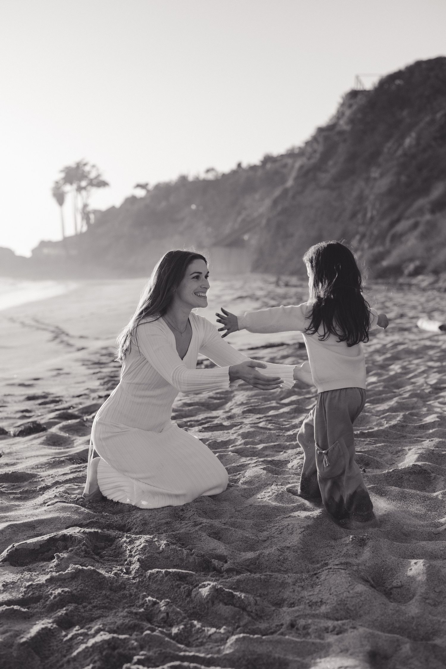A tender moment captured in black and white on a beach with cliffs and palm trees in the background.