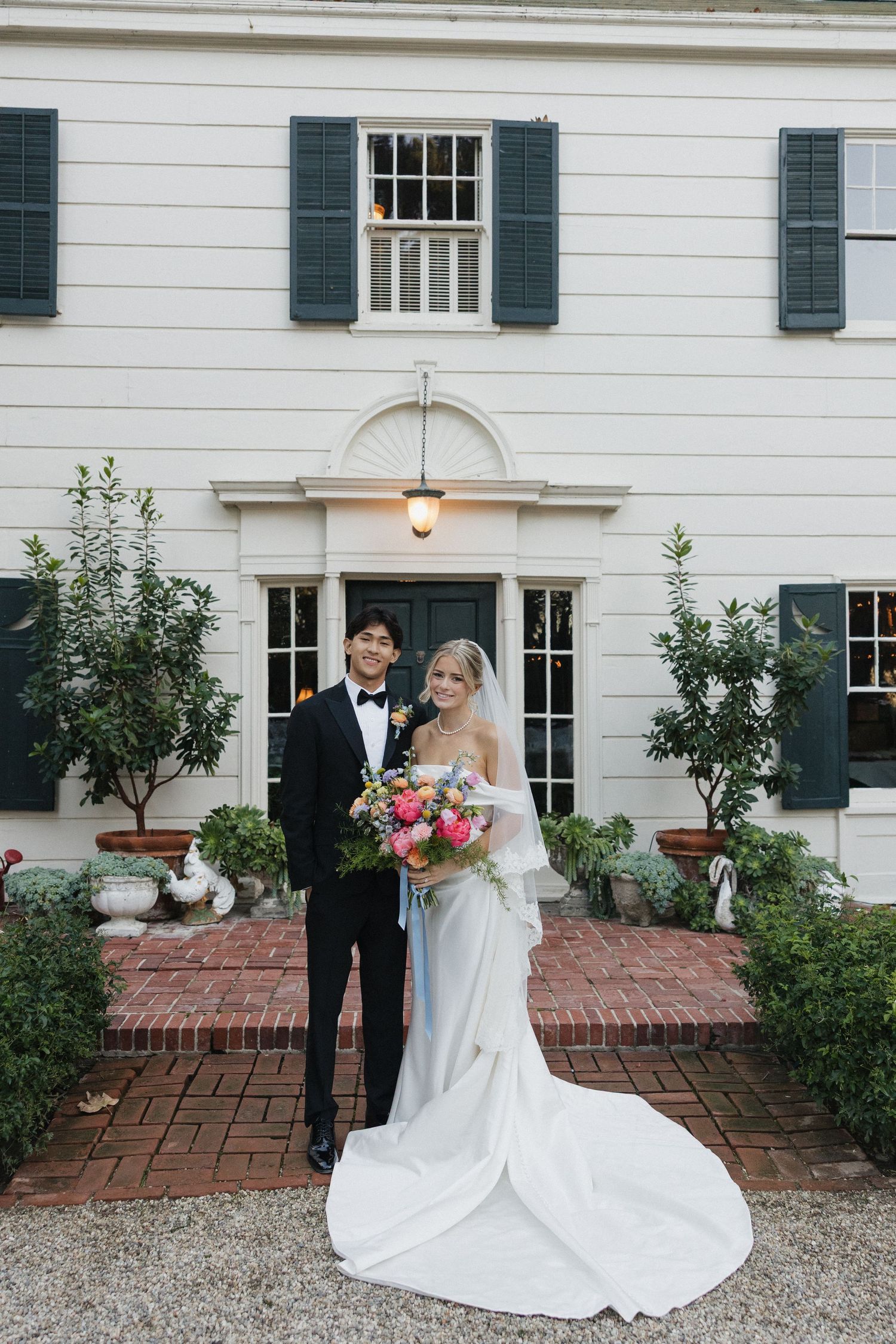 A classic white colonial house with black shutters serves as an elegant backdrop for wedding portraits.