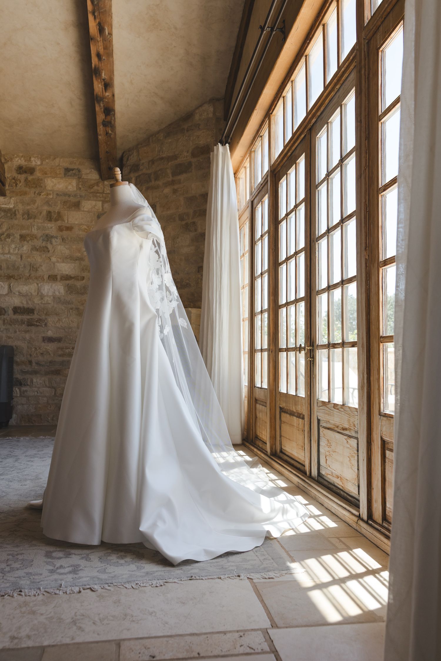 An elegant white wedding dress with a cape hangs by a sunlit window in a stone-walled room.