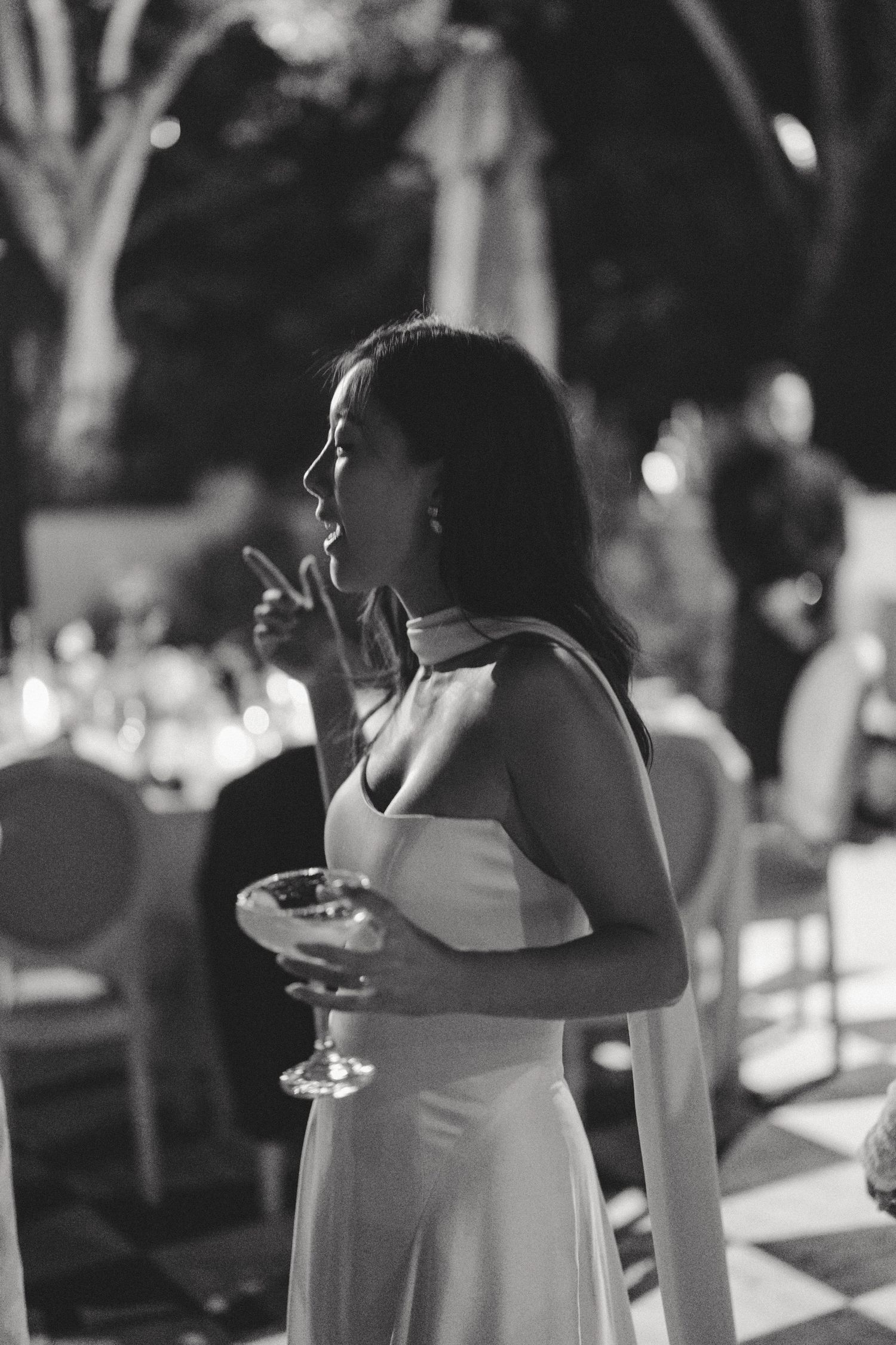 A guest in a white dress holds a cocktail glass during an evening outdoor reception.