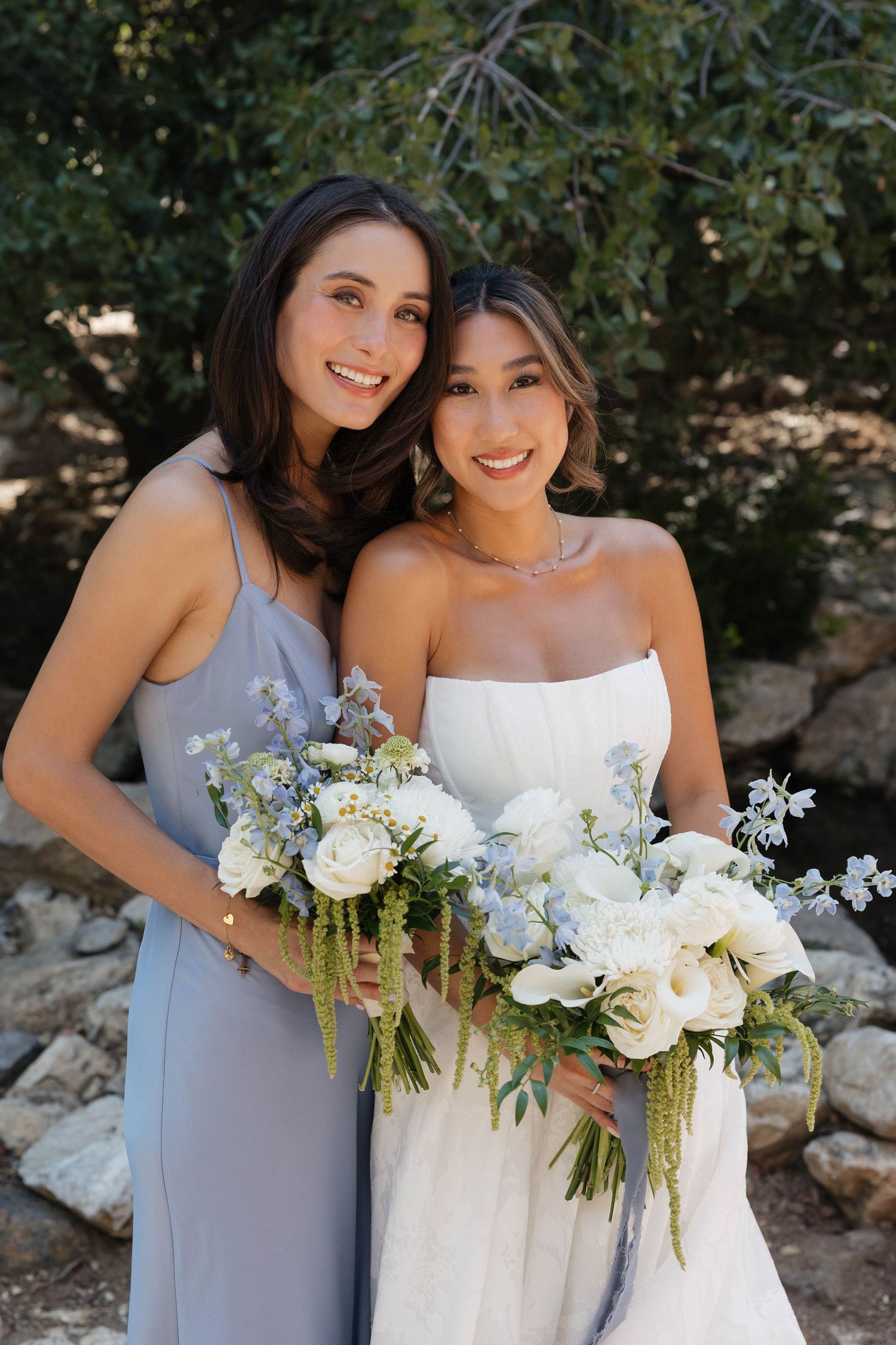 Bride in white gown and bridesmaid in gray dress holding beautiful white bouquets outdoors.