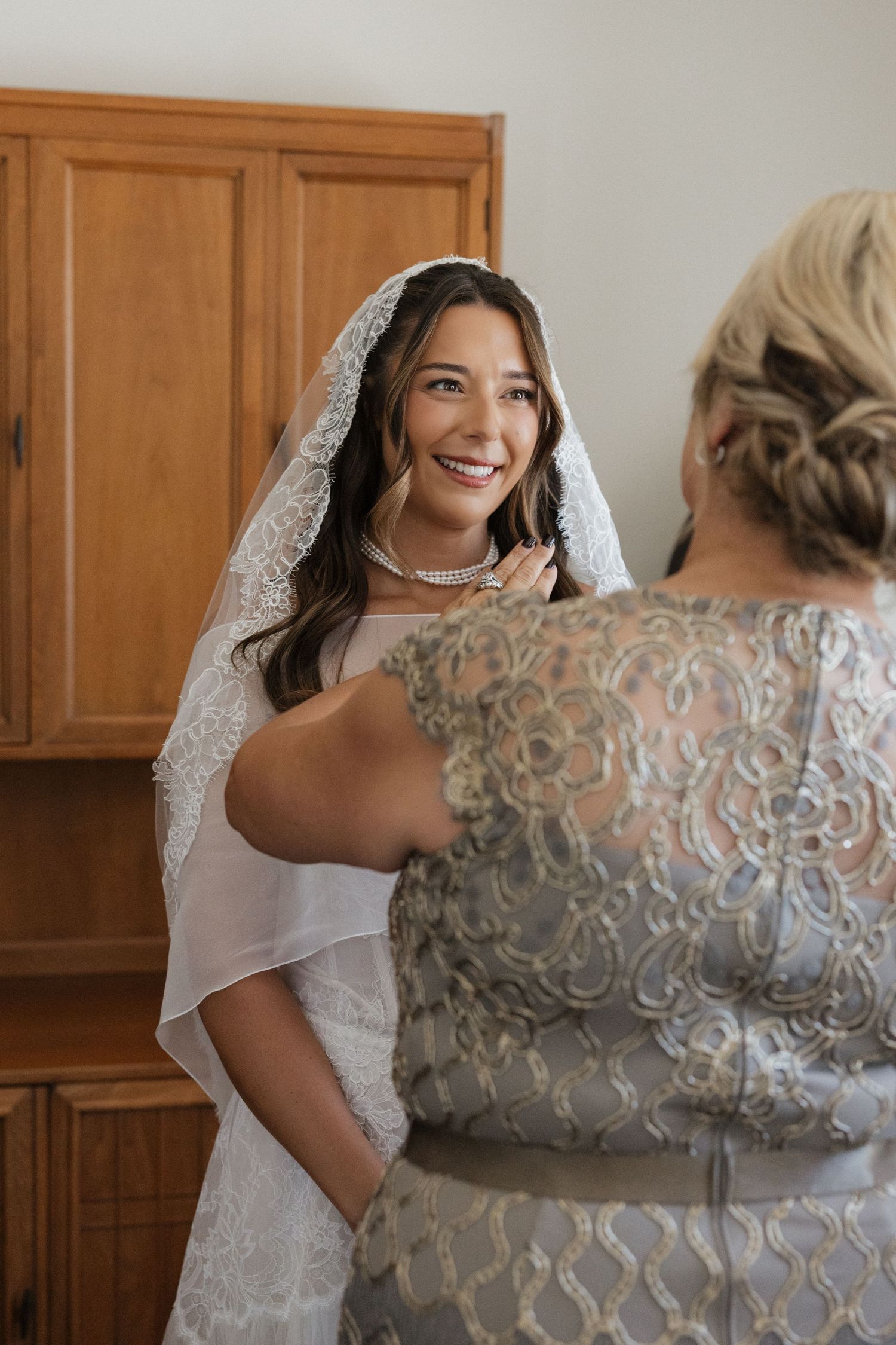 Smiling bride in lace veil gets necklace fastened by woman in silver embroidered gown before wedding ceremony.