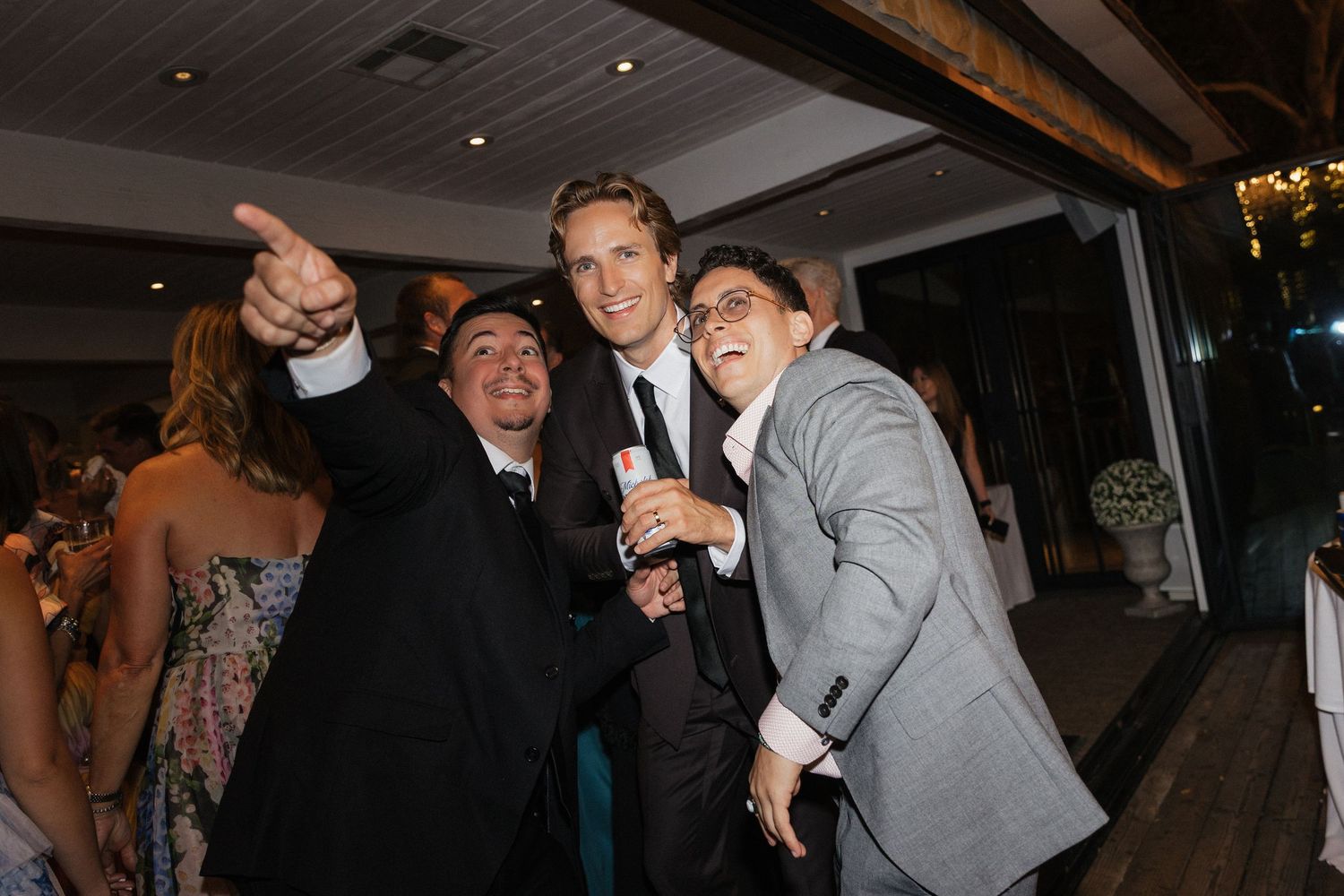 Three men smiling and pointing at a lively wedding reception party.