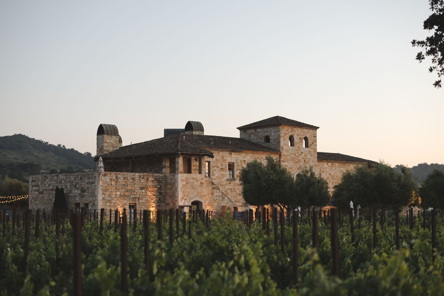 Vineyard estate with stone buildings and mountains in background at sunset.
