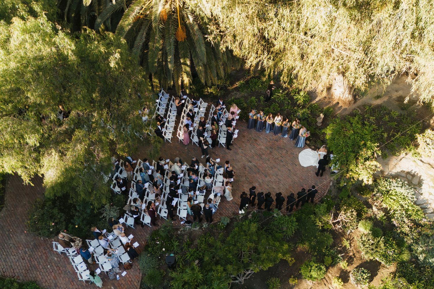 Aerial view of an outdoor wedding ceremony with guests arranged in rows among trees and natural scenery.