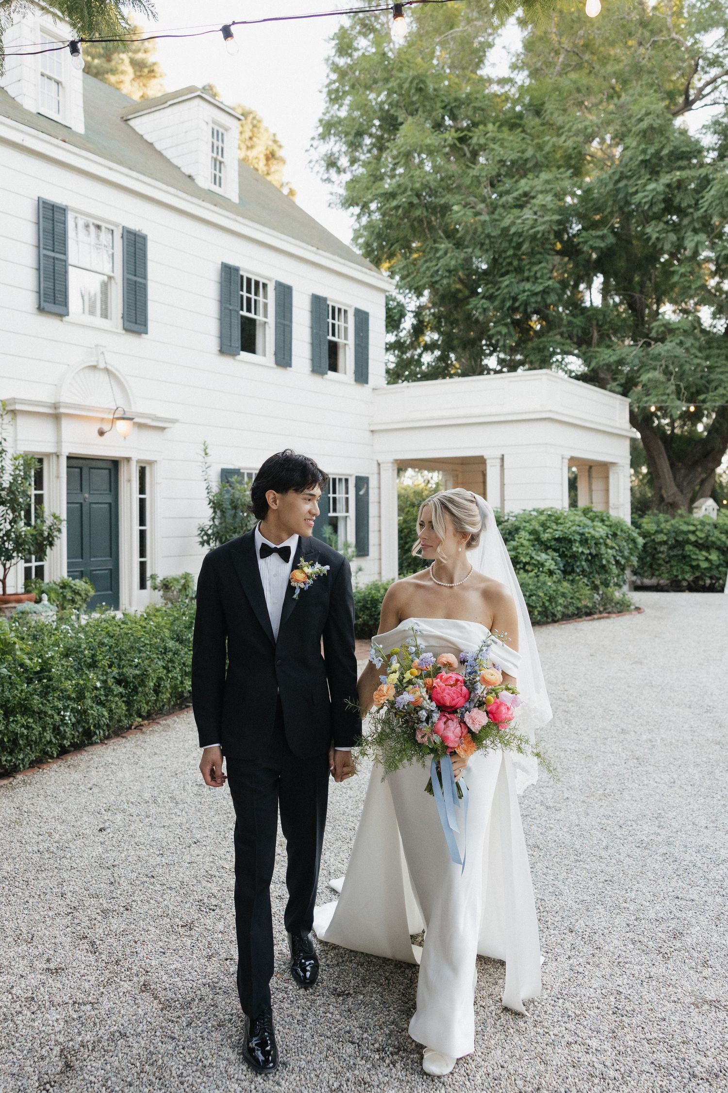 A wedding couple walks along a white gravel path beside a classic colonial-style mansion.