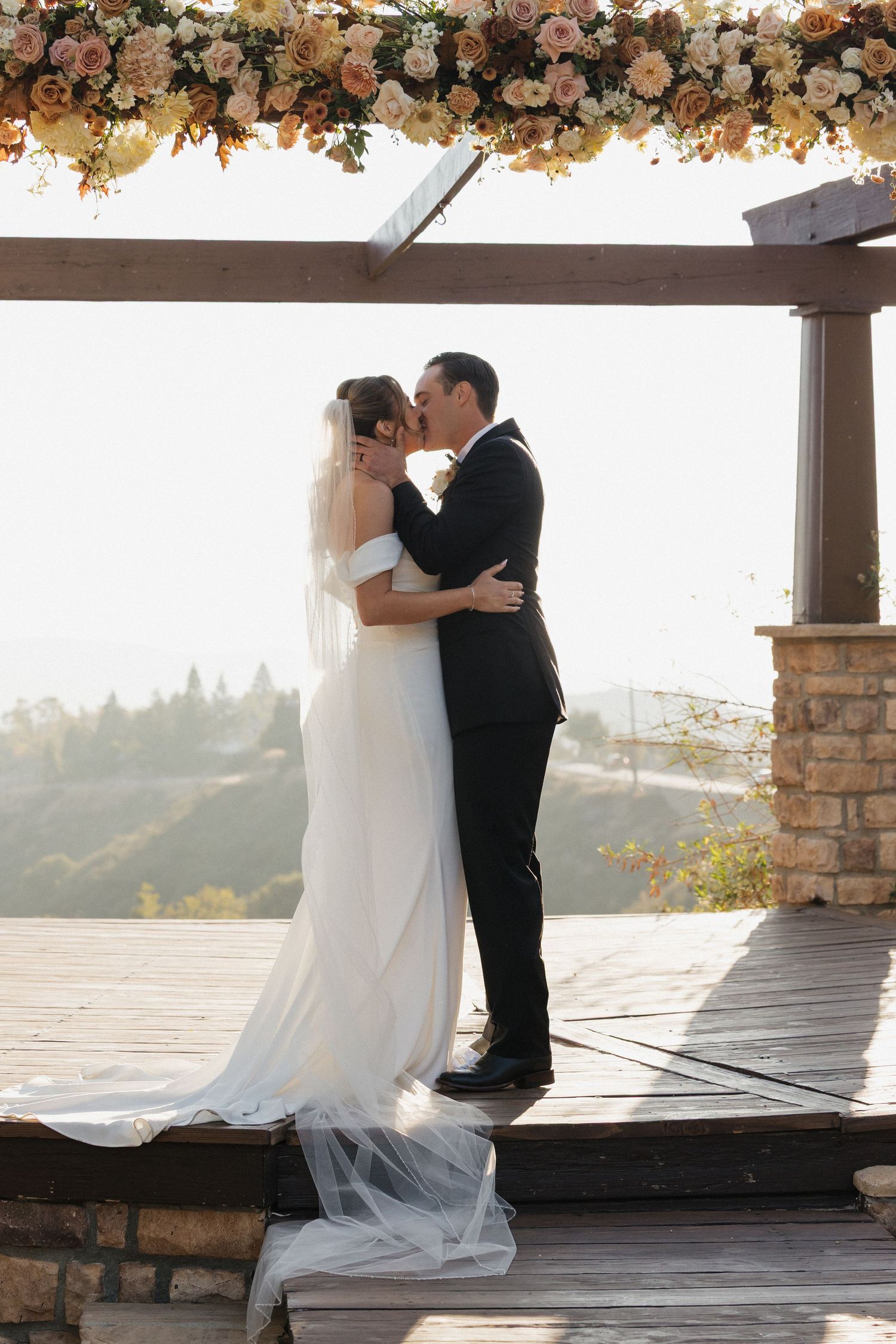 Wedding couple embracing under a floral-decorated pergola during golden hour.