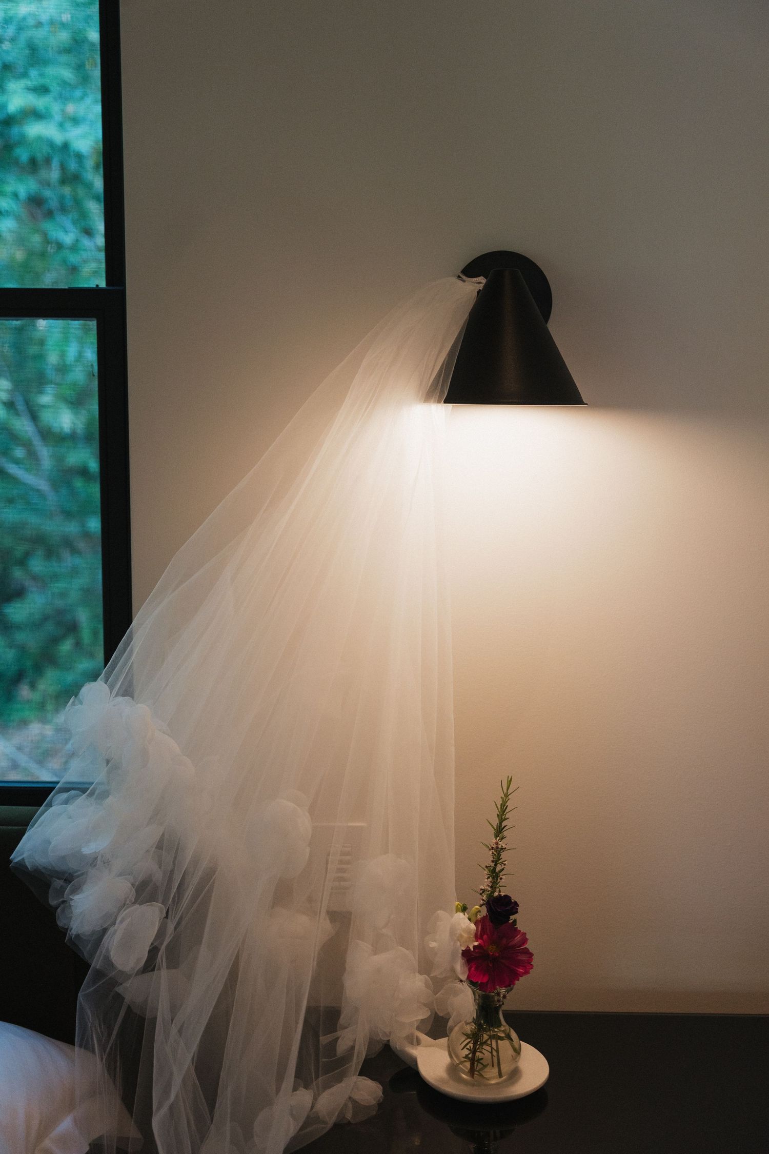 A white wedding veil and red flower arrangement sit beneath a desk lamp on a table.