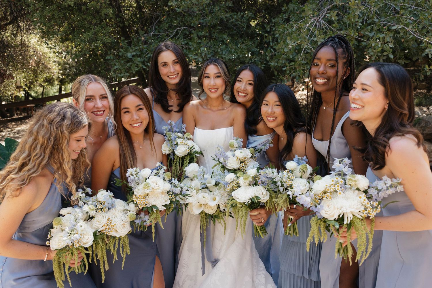 Bridesmaids in blue dresses hold white bouquets while sharing joyful moment outdoors.