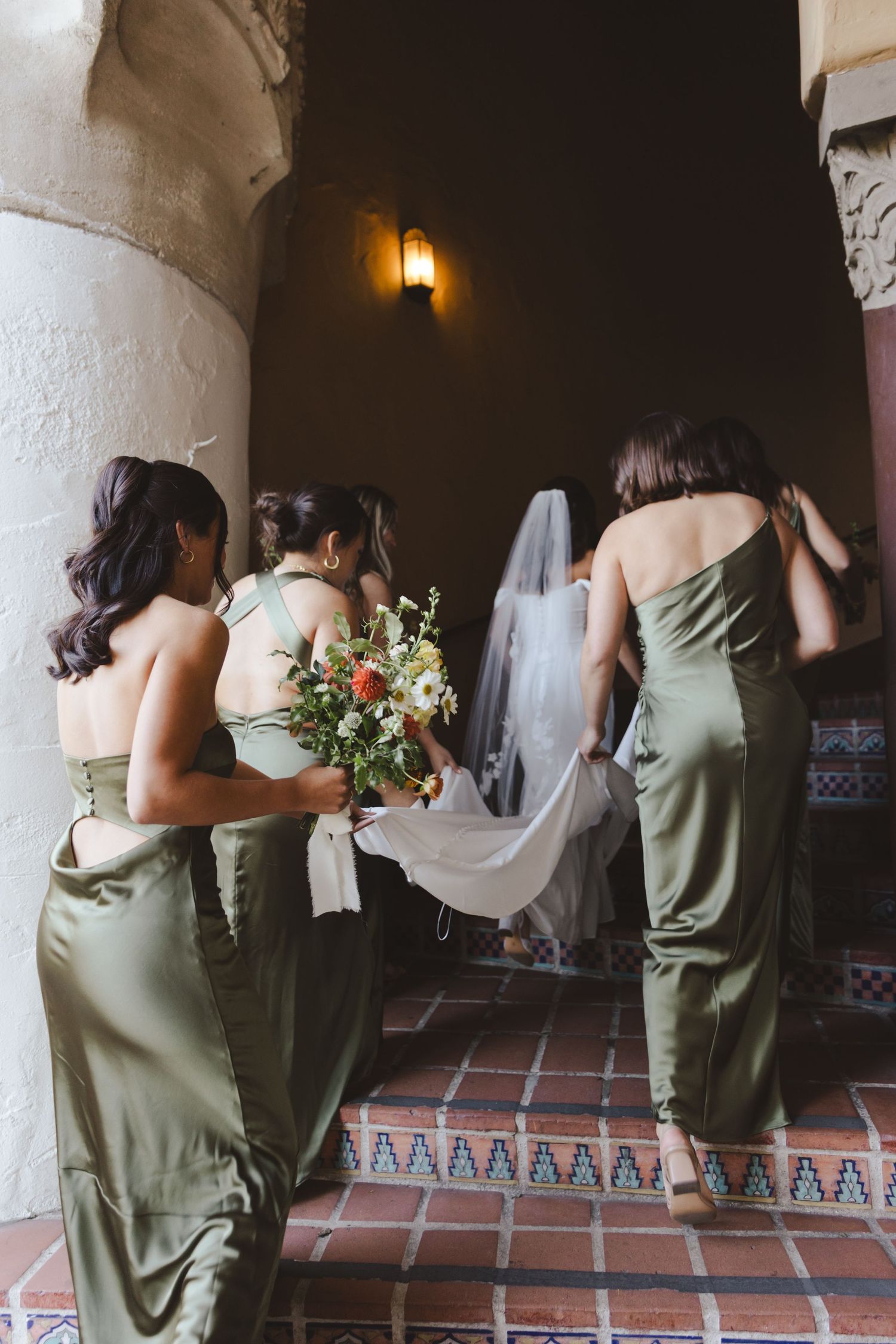 Bridesmaids in sage green dresses walk up tiled stairs carrying bouquets and helping with the bride's veil.