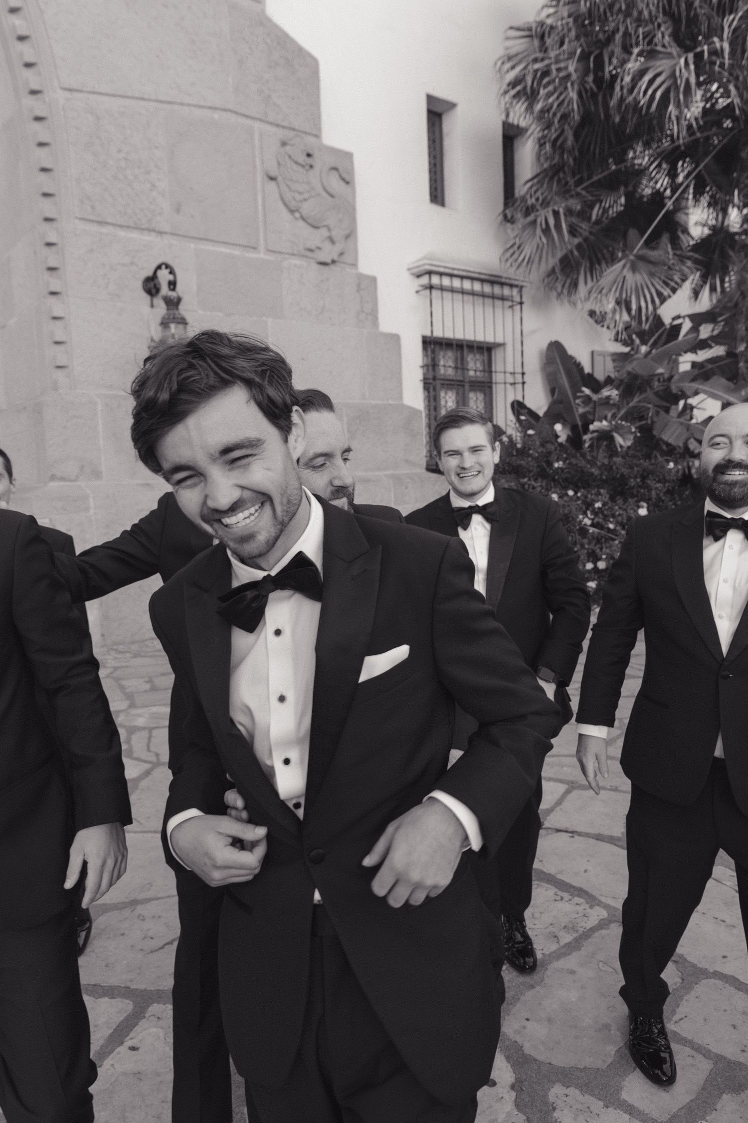 Groomsmen in classic black tuxedos share laughter during a candid black and white wedding photo.