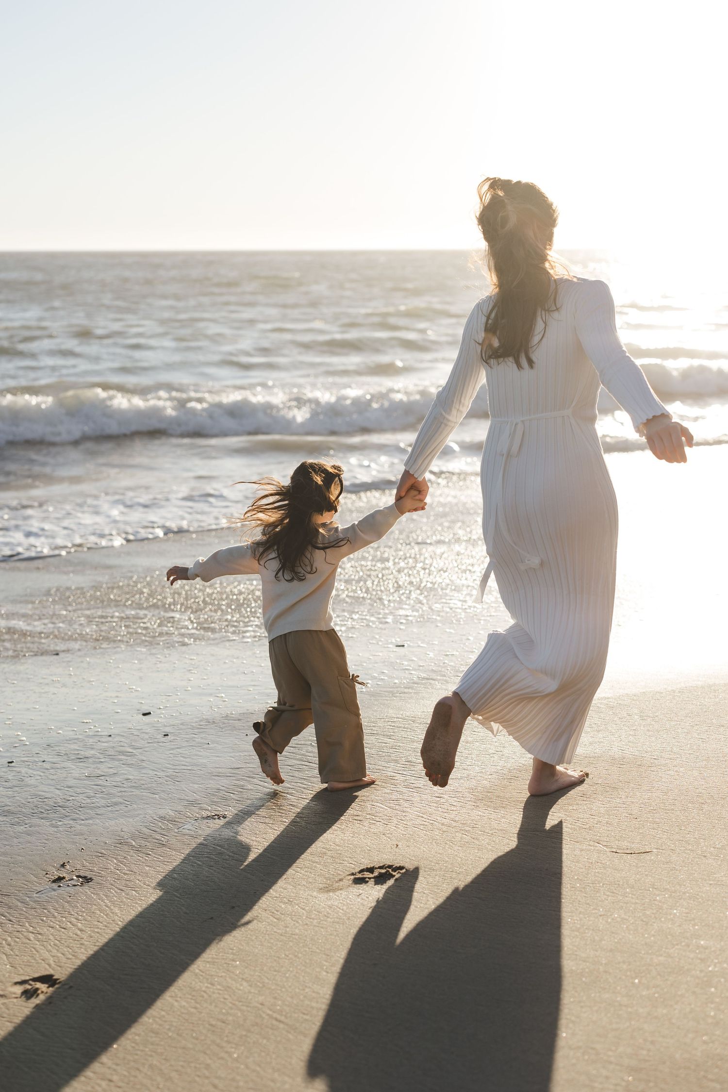 A parent and child holding hands while walking and playing on a sunny beach at sunset as waves roll in.