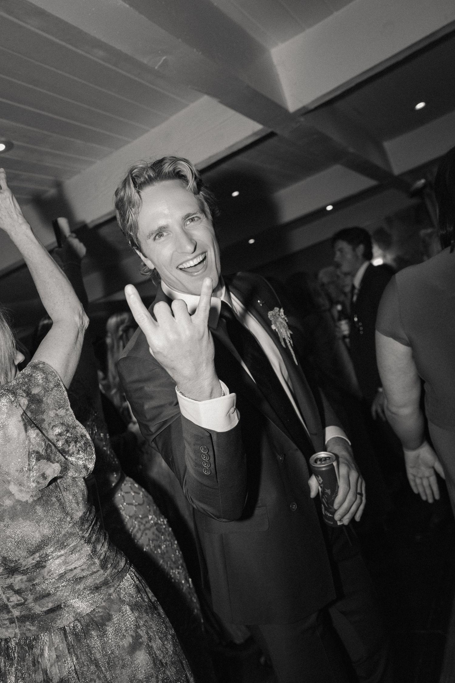 Black and white photo of grinning groom making rock sign on wedding dance floor holding a drink.