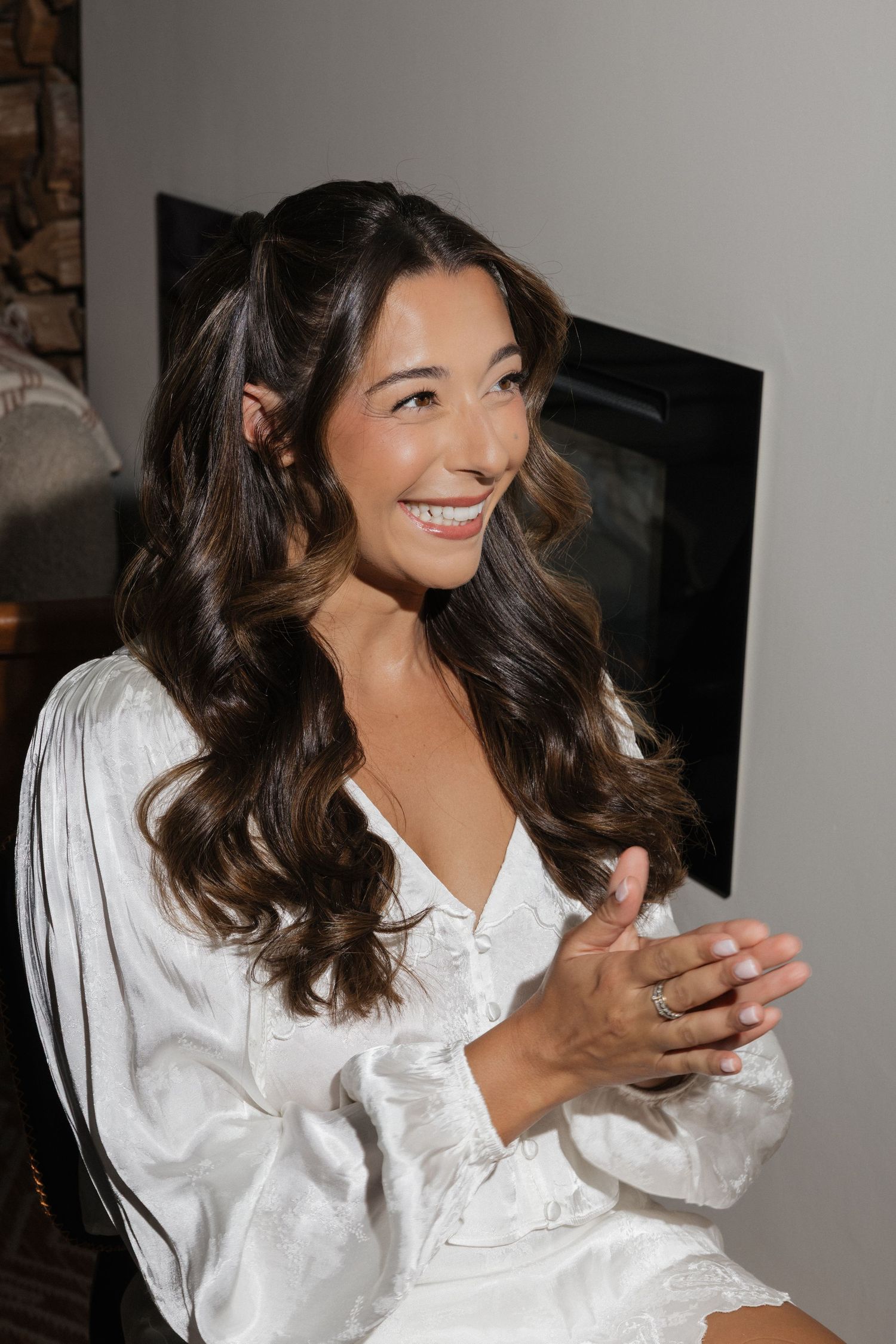Smiling bride in white satin robe with long wavy brunette hair clasping hands near fireplace.