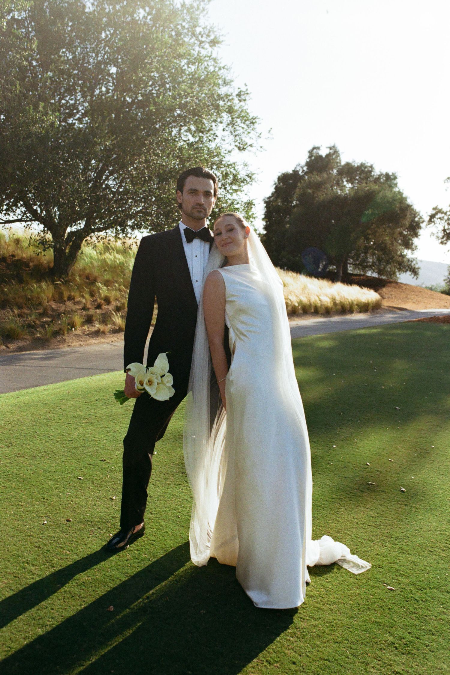 Bride in white gown and veil stands with groom in black tuxedo on green lawn surrounded by trees.