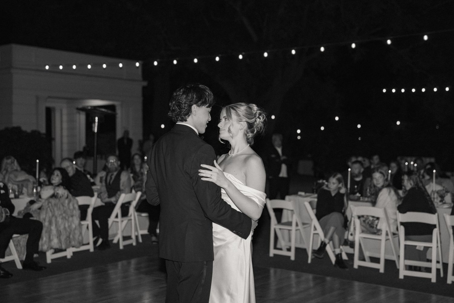 String lights twinkle above an outdoor wedding dance floor as guests celebrate in the evening.