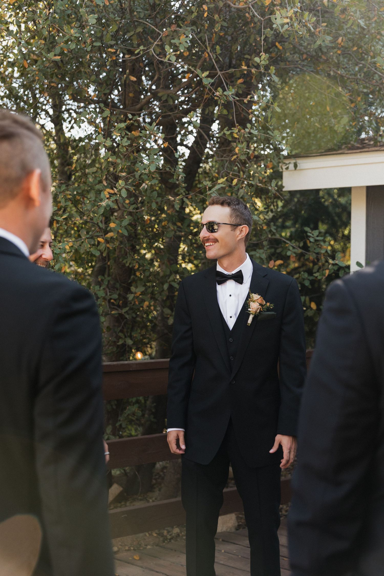 A groom in a classic black tuxedo and sunglasses stands outdoors during a wedding ceremony.