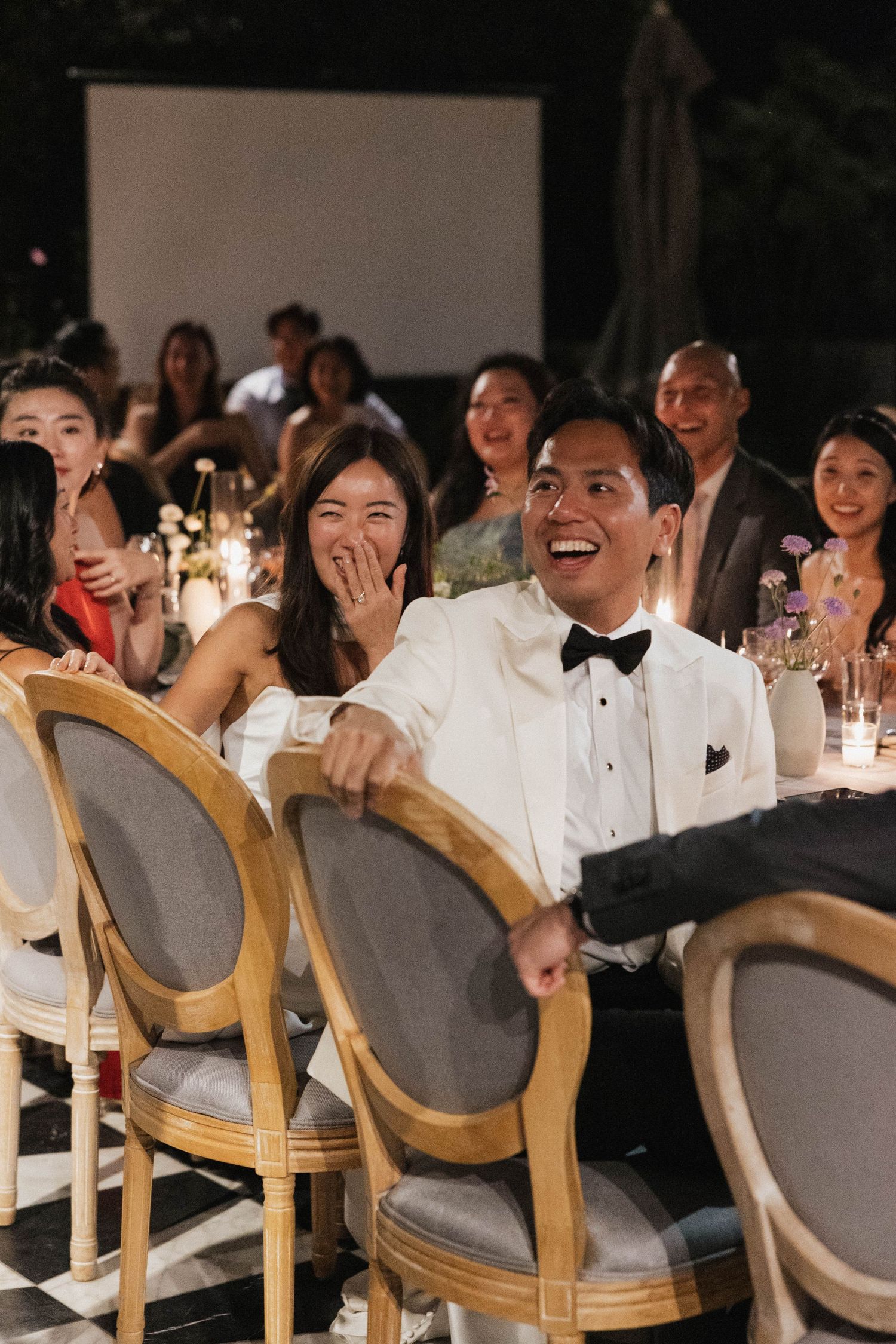 Wedding guests laughing during reception while seated in elegant wooden chairs.
